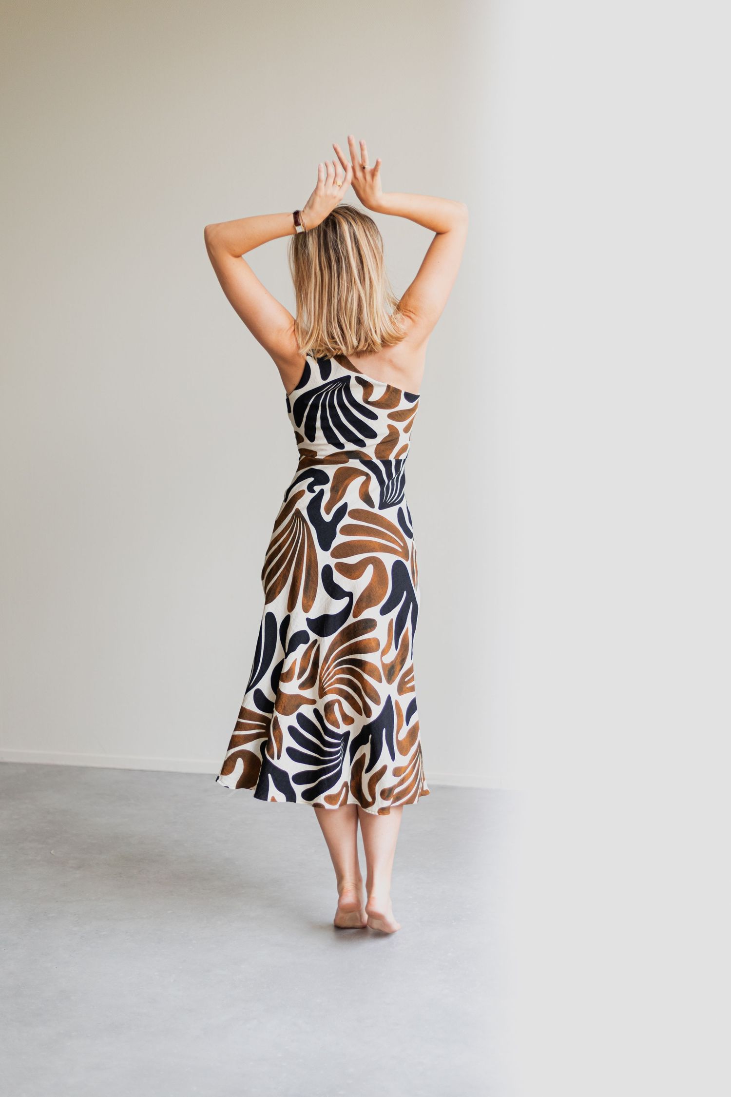Woman in abstract print midi dress with arms raised, viewed from behind, standing against neutral wall.