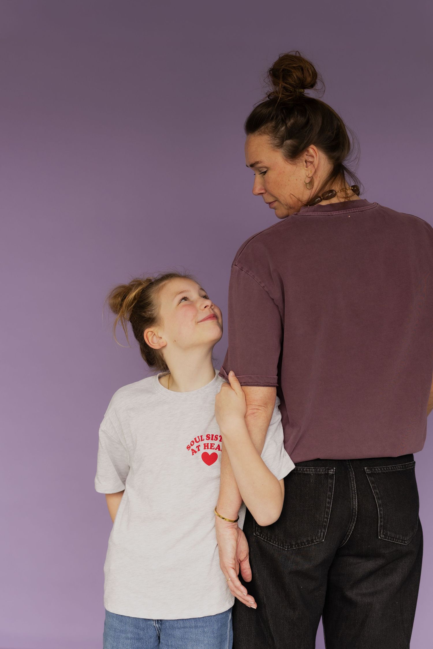 A young girl and an older woman in matching bun hairstyles share a tender moment against a purple background.