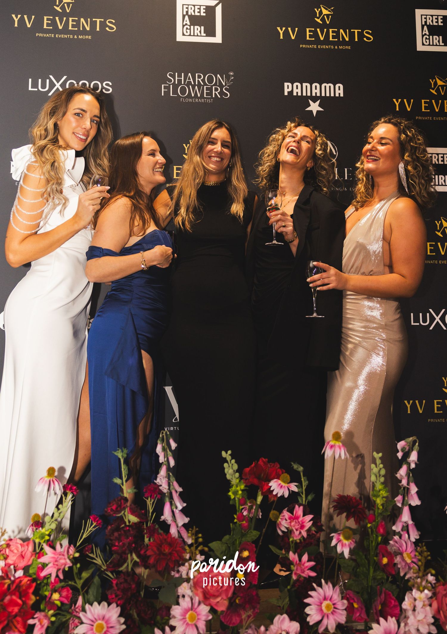 A group of elegantly dressed women laugh and celebrate at a formal event with floral decorations and branded backdrops.