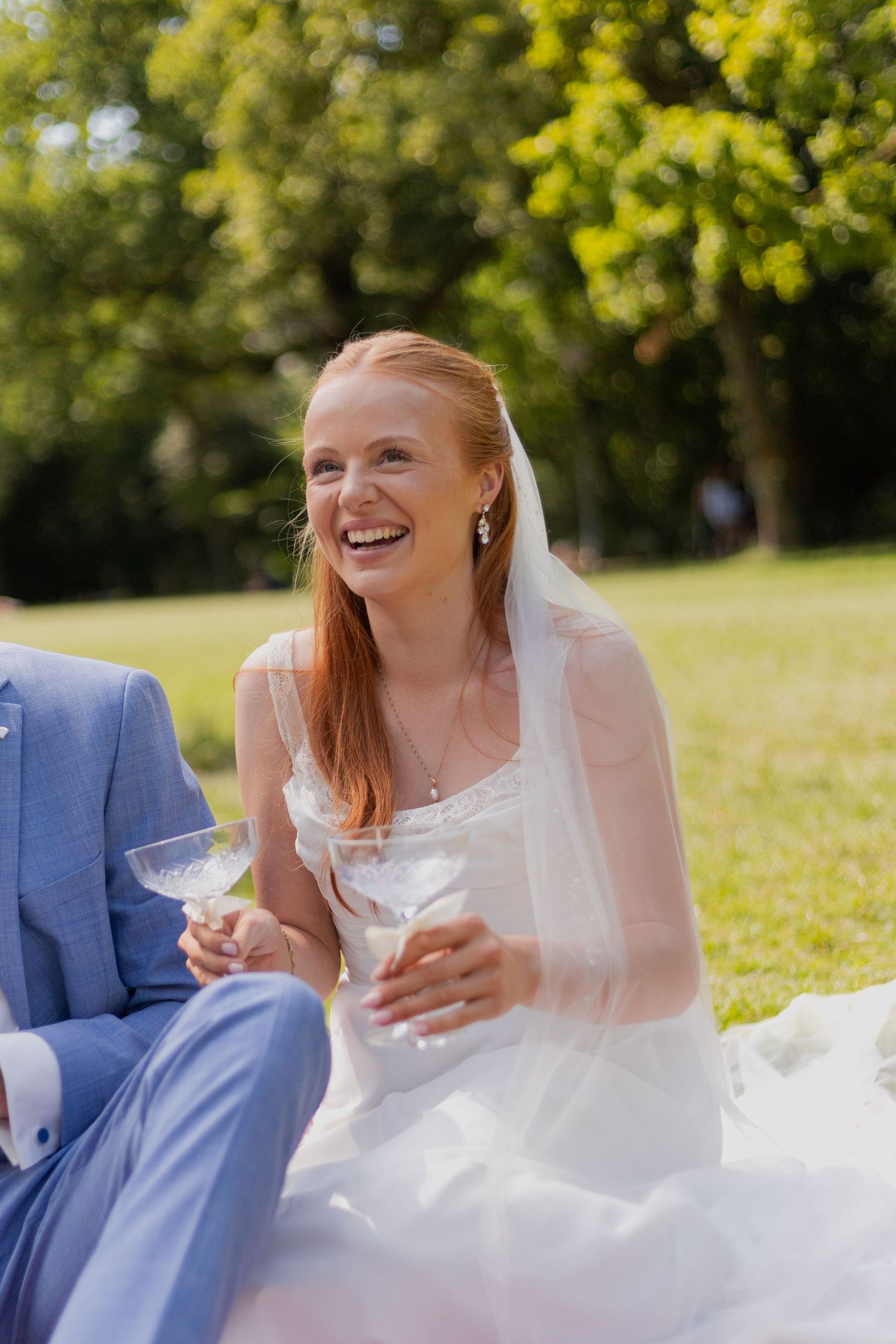 A couple sits on grass sharing drinks and laughter during their wedding celebration.