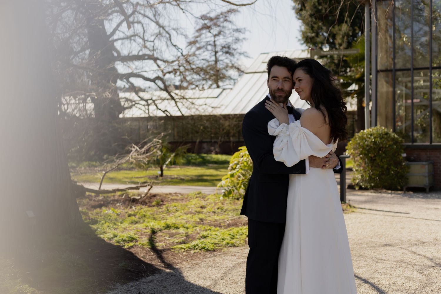 Couple in wedding attire walking along a sunlit garden path.