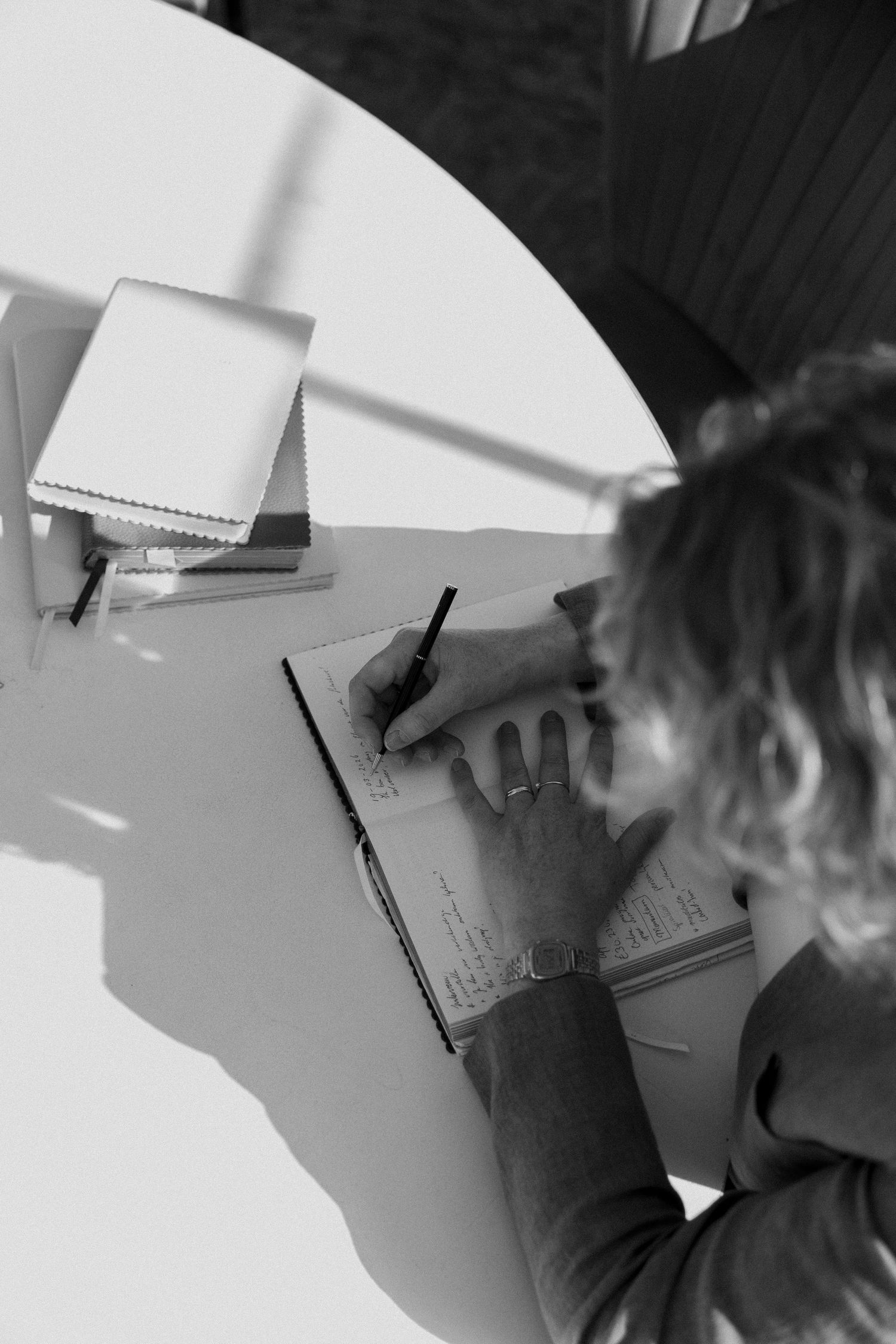 Black and white overhead photo of a woman writing in a notebook at a round white table outdoors.