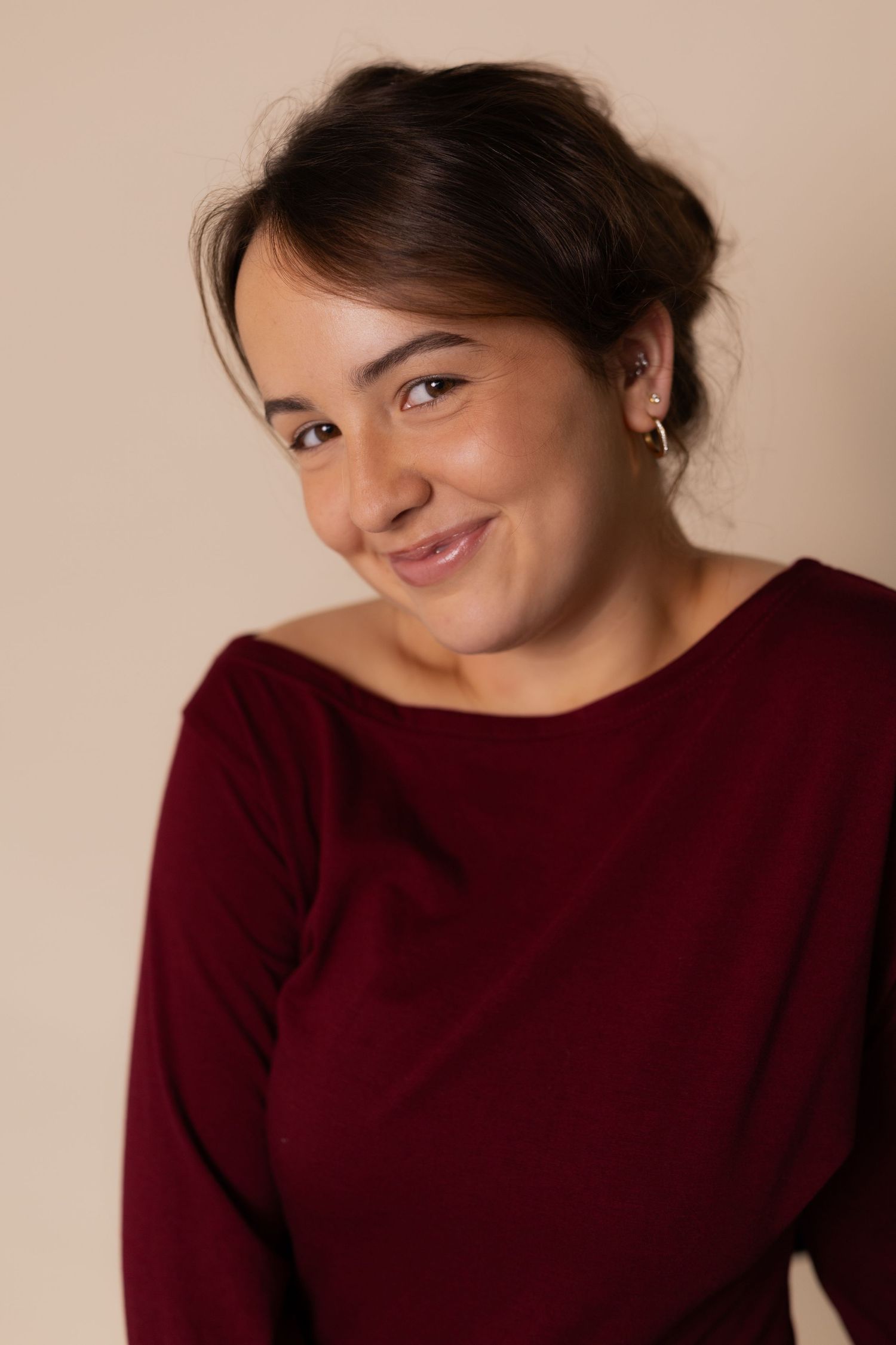 Young woman with dark updo hair smiling warmly, wearing a burgundy top against a beige background.