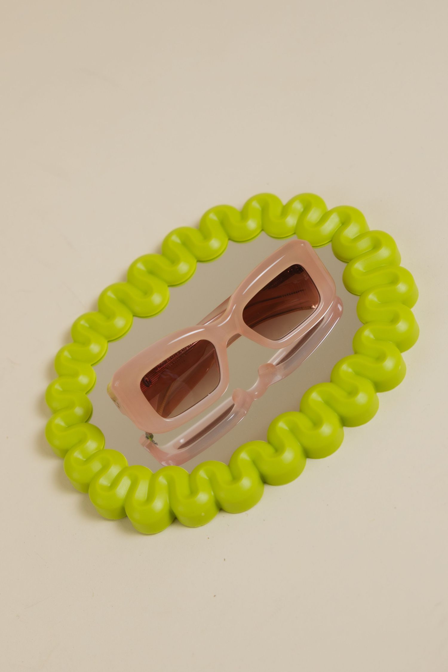 Pink rectangular sunglasses displayed on a reflective oval mirror framed by a bright lime green bubble border.
