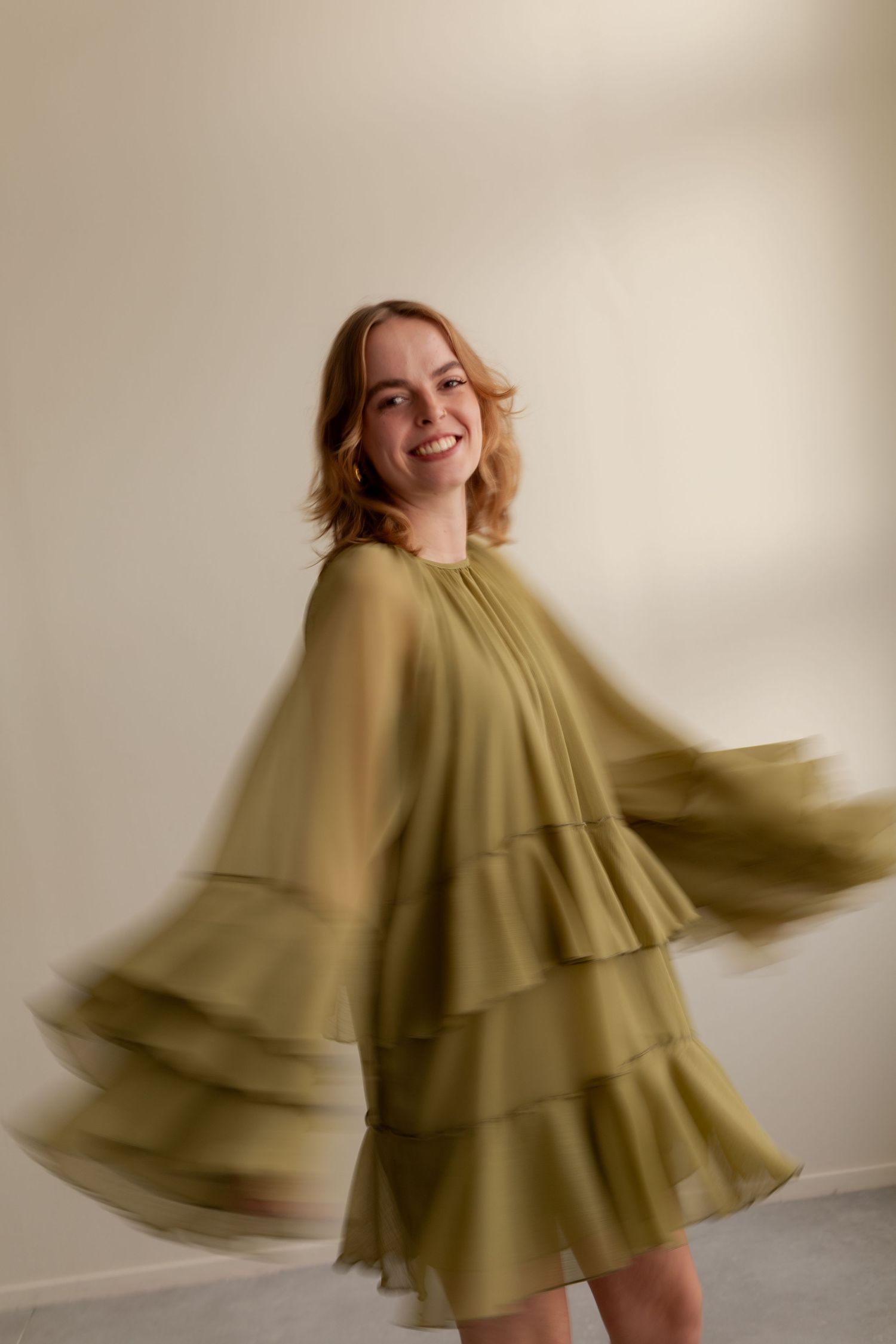 A person in an olive green tiered dress spins and twirls in a sequence of motion blur photos against a white wall.