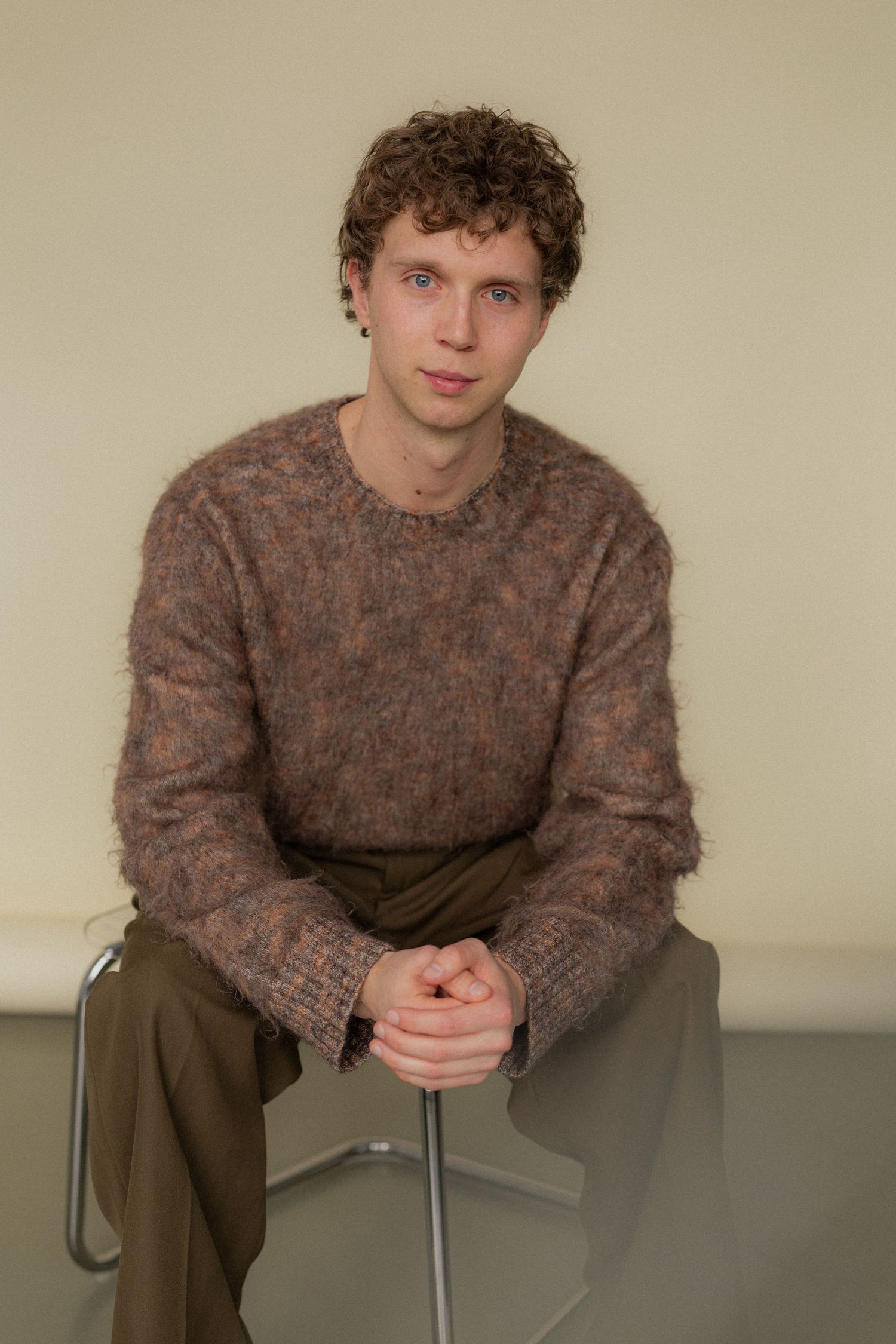 A person in a brown knit sweater sits casually on a chair against a neutral wall.