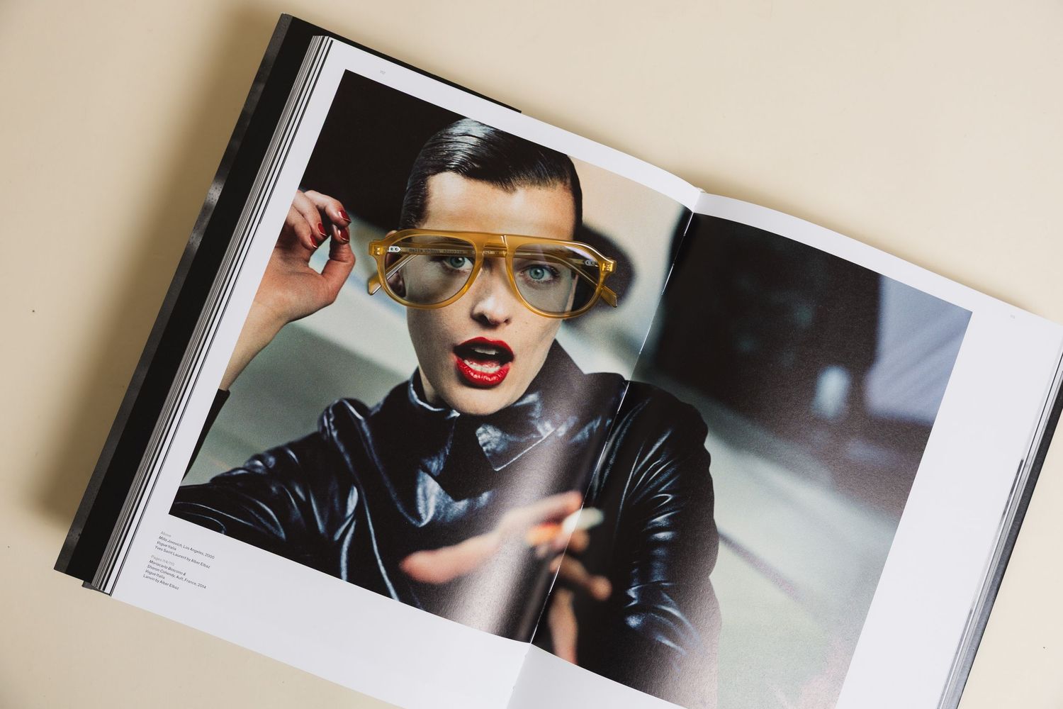 Fashion editorial book open to a page featuring a model in black leather jacket wearing bold yellow-framed glasses.