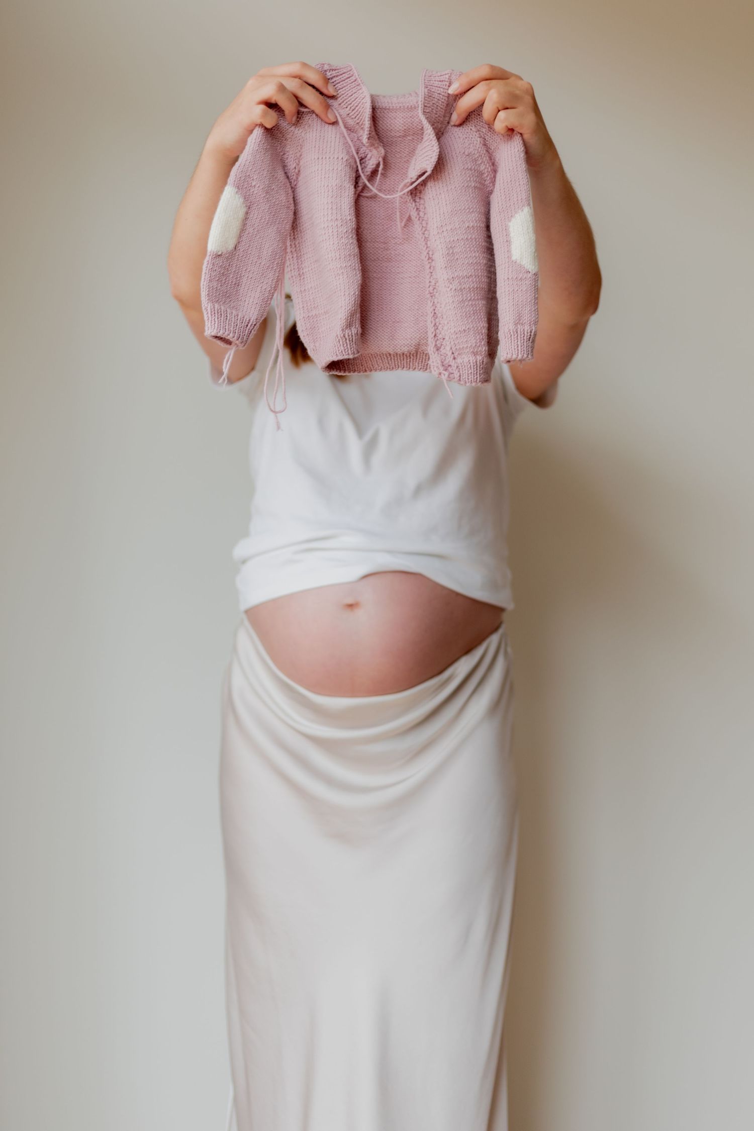 A pregnant person in white dress holds up a small pink baby cardigan against a neutral wall.