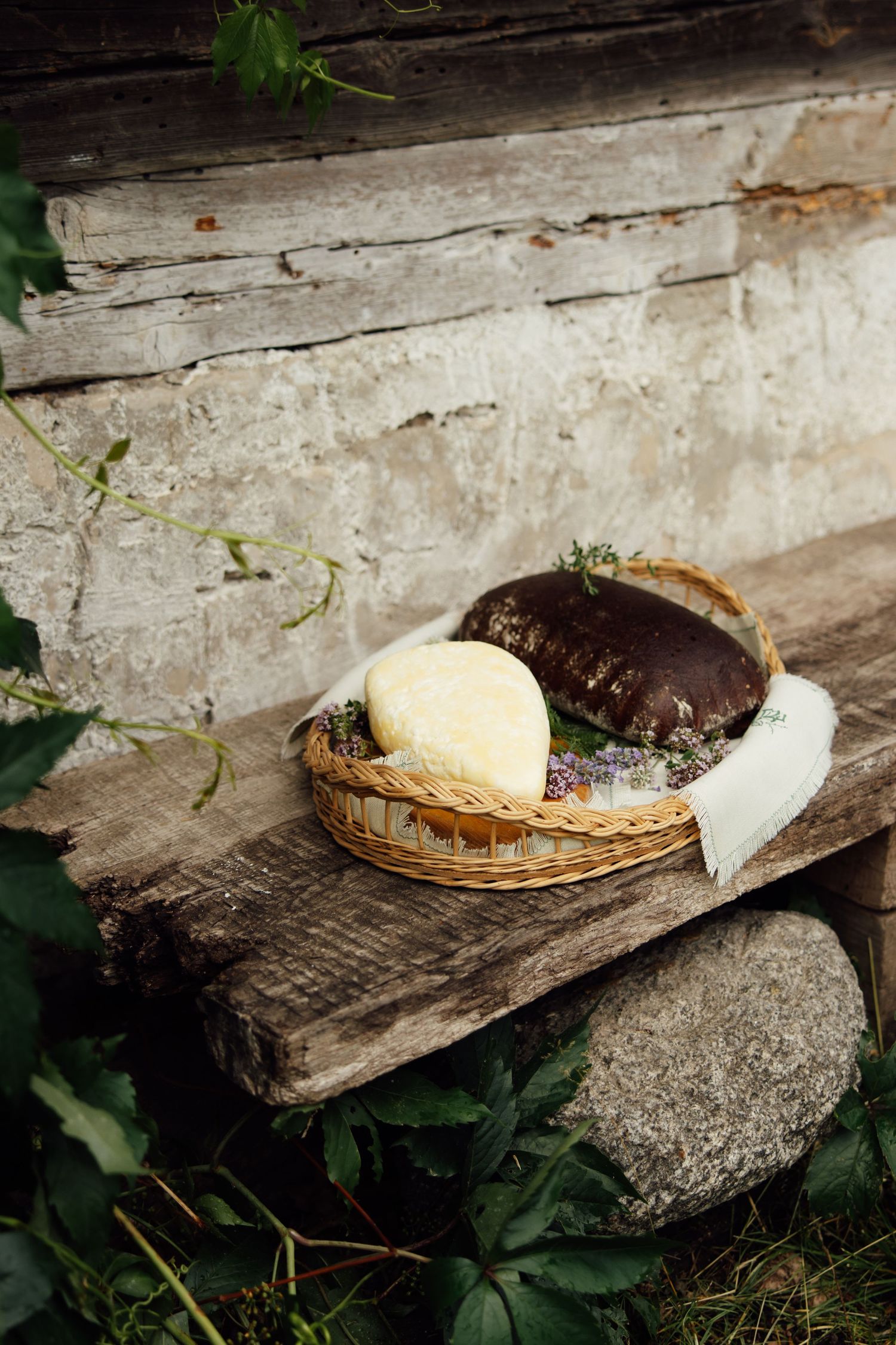 Rustic wicker basket with artisan cheese and dark bread on weathered wooden bench outdoors.