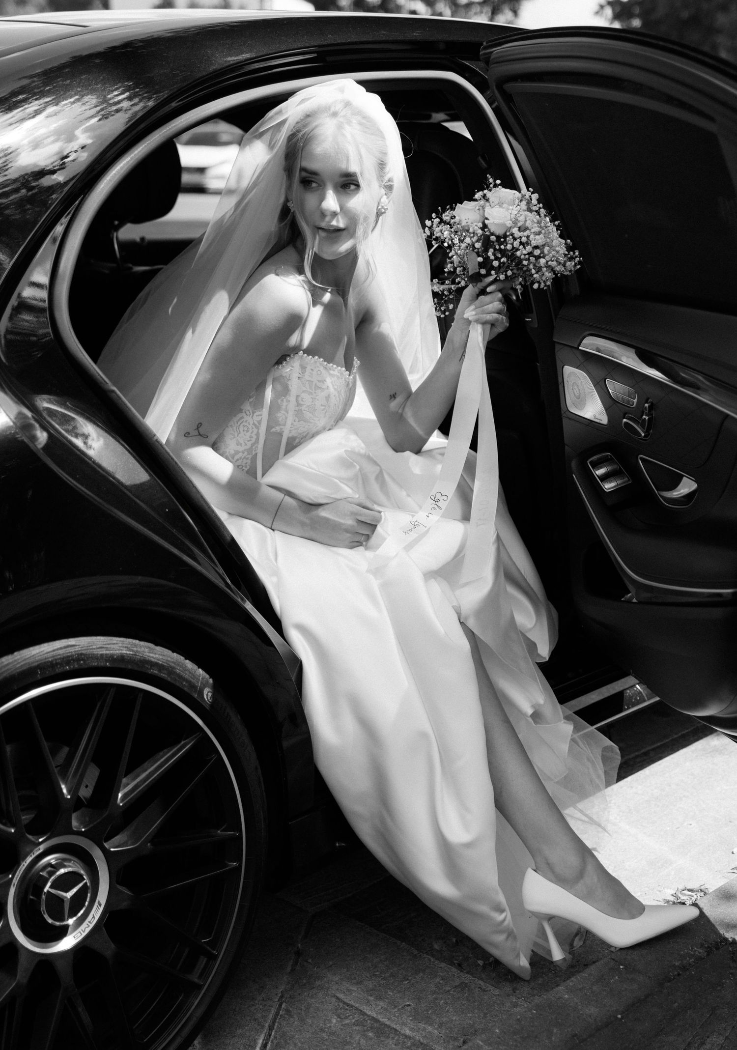 Bride in white gown and veil holding bouquet, stepping out of a Mercedes car, black and white wedding photo.