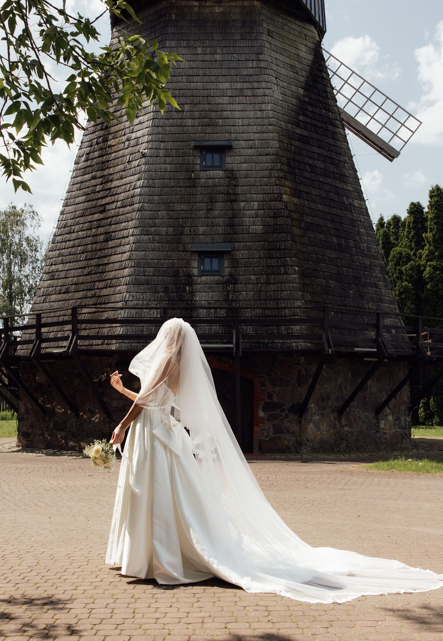 Bride in white gown and veil stands before a historic wooden windmill on a brick pathway.