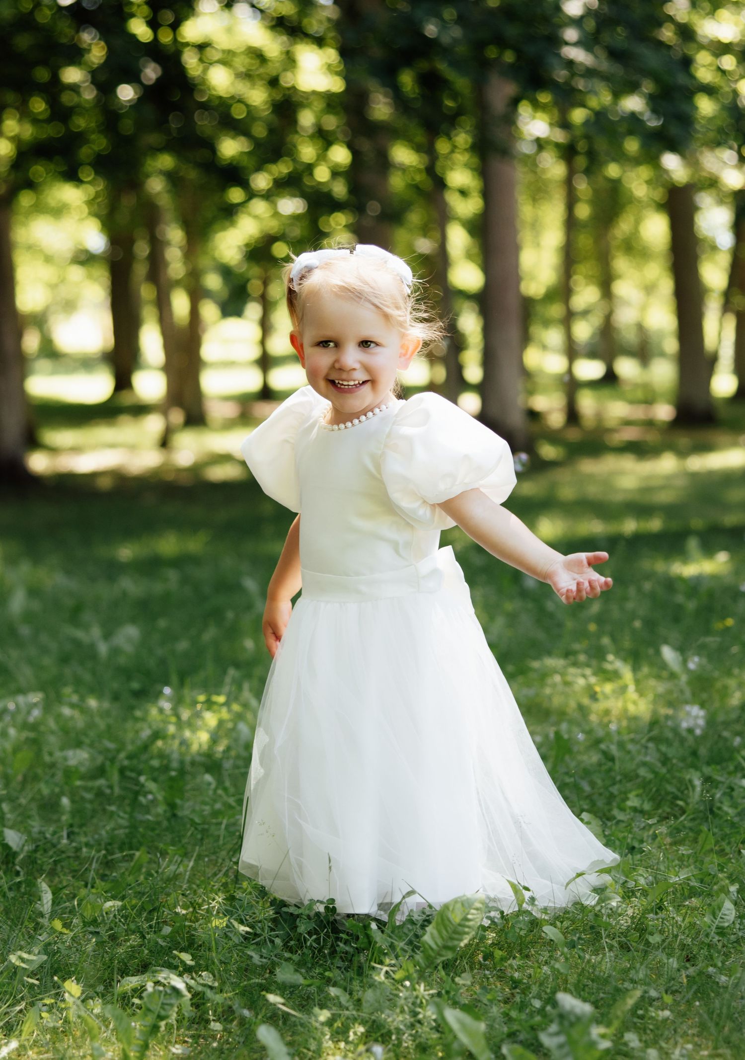 Smiling toddler girl in white flower girl dress with puff sleeves and tulle skirt playing outdoors in green park.