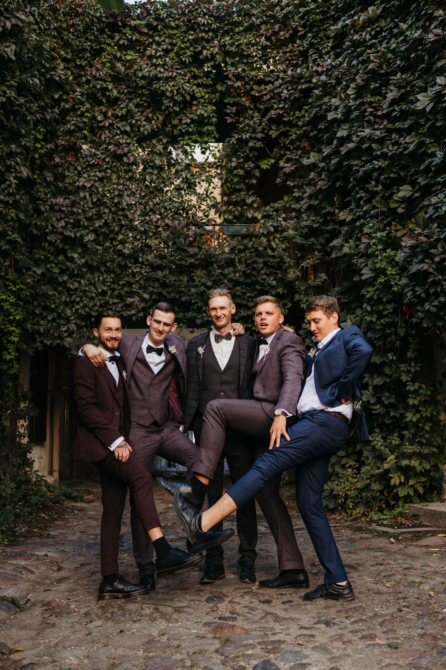 Groomsmen in suits pose casually against an ivy-covered wall for a fun group photo.