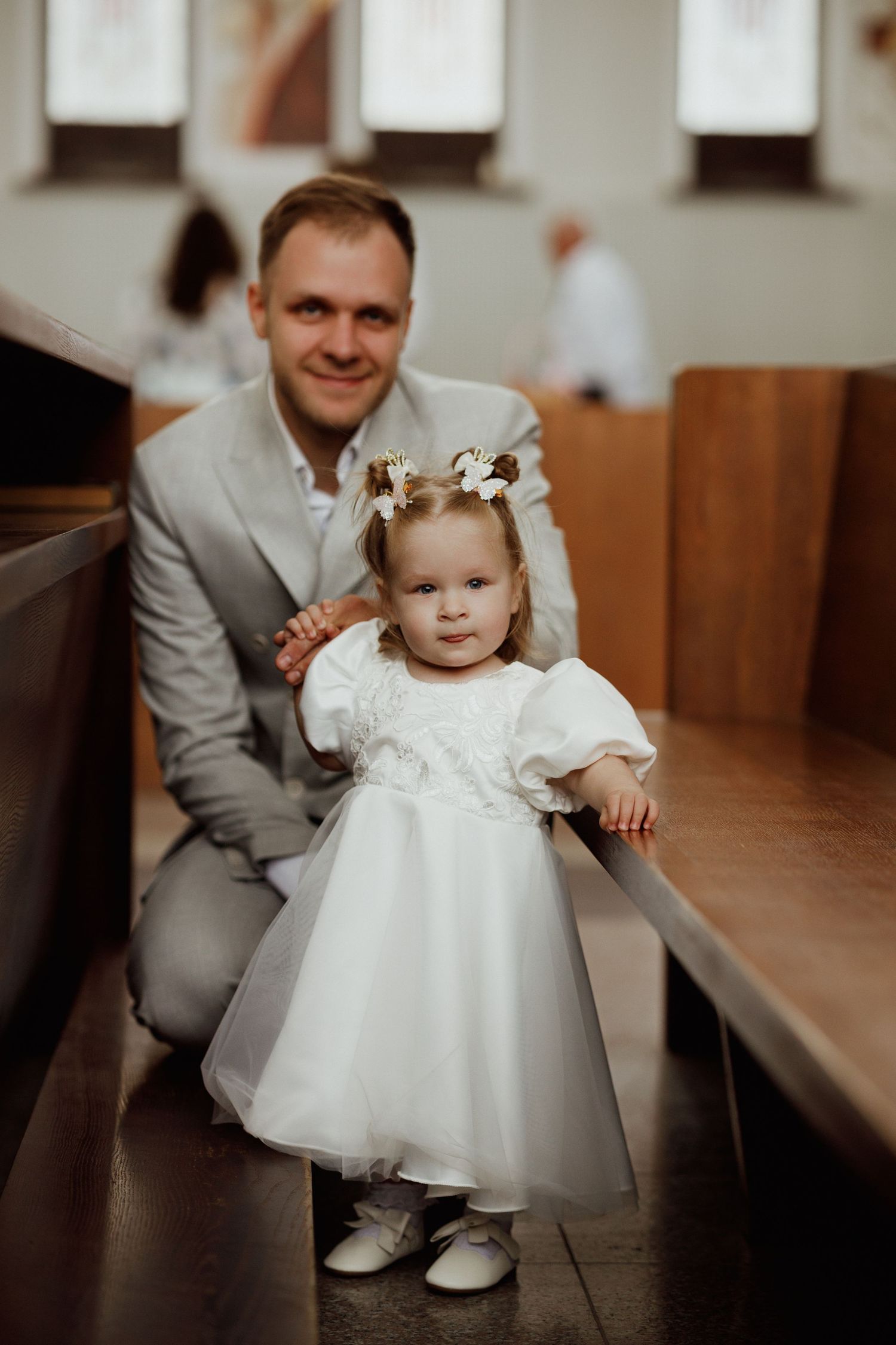 A small child in a white christening dress sits on a wooden church pew while an adult in gray looks on.