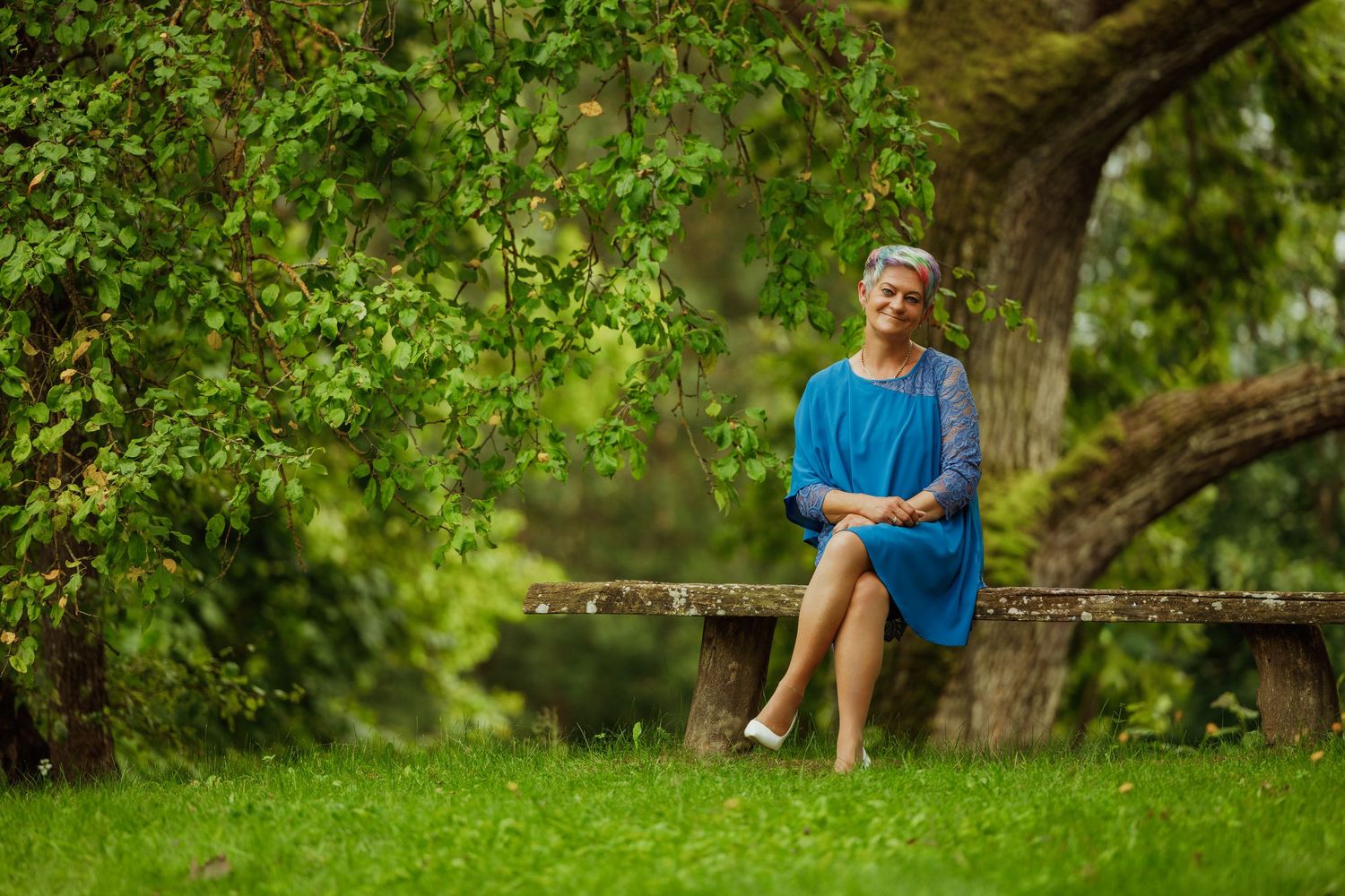 A person wearing blue sits casually on a wooden bench surrounded by lush green foliage in a garden.