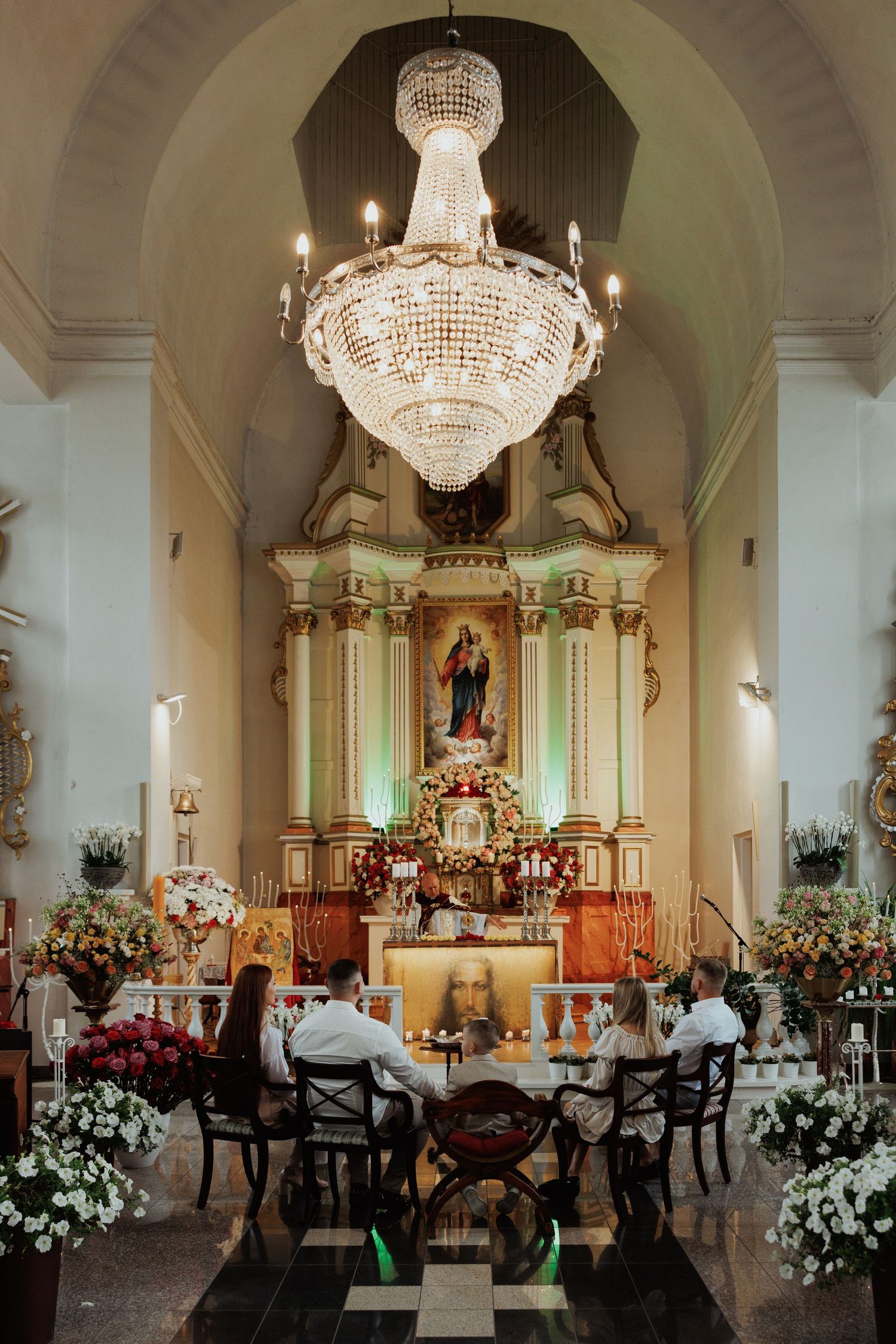Elegant church interior with crystal chandelier, ornate altar, and white floral decorations during a wedding ceremony.