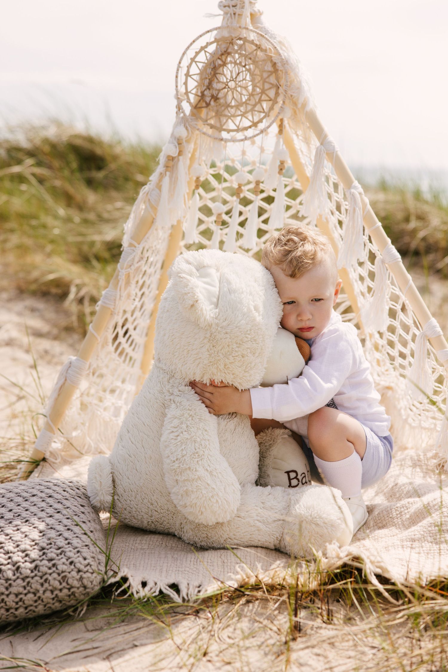 A dreamy beach scene with a large teddy bear and a lace teepee setup on the sand.