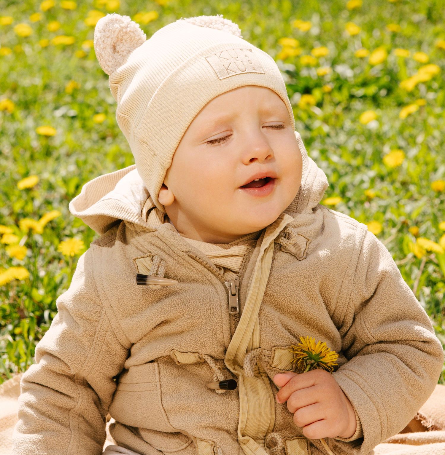 A small child wearing a beige winter coat and knit hat sits in a field of yellow dandelions on a sunny day.