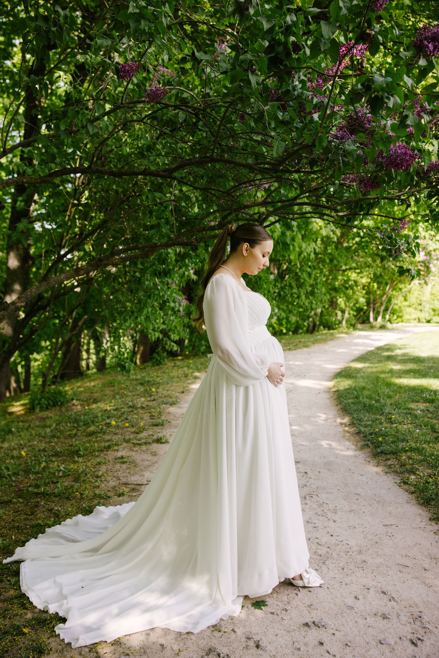 An elegant wedding dress with flowing train photographed on a garden path beneath blooming lilac trees.