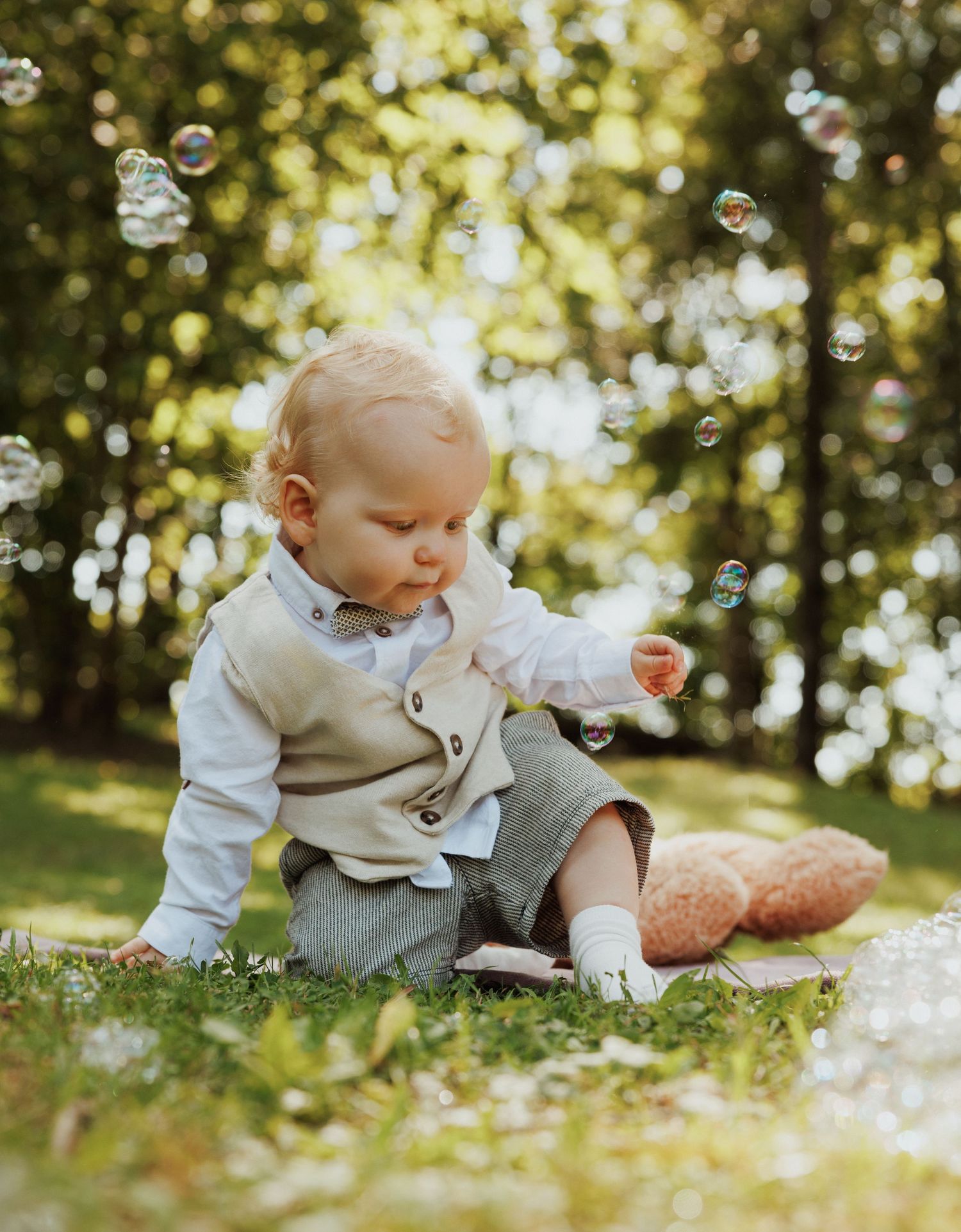 Baby in beige overalls plays with bubbles on grass in a sunlit garden during an outdoor photo session.