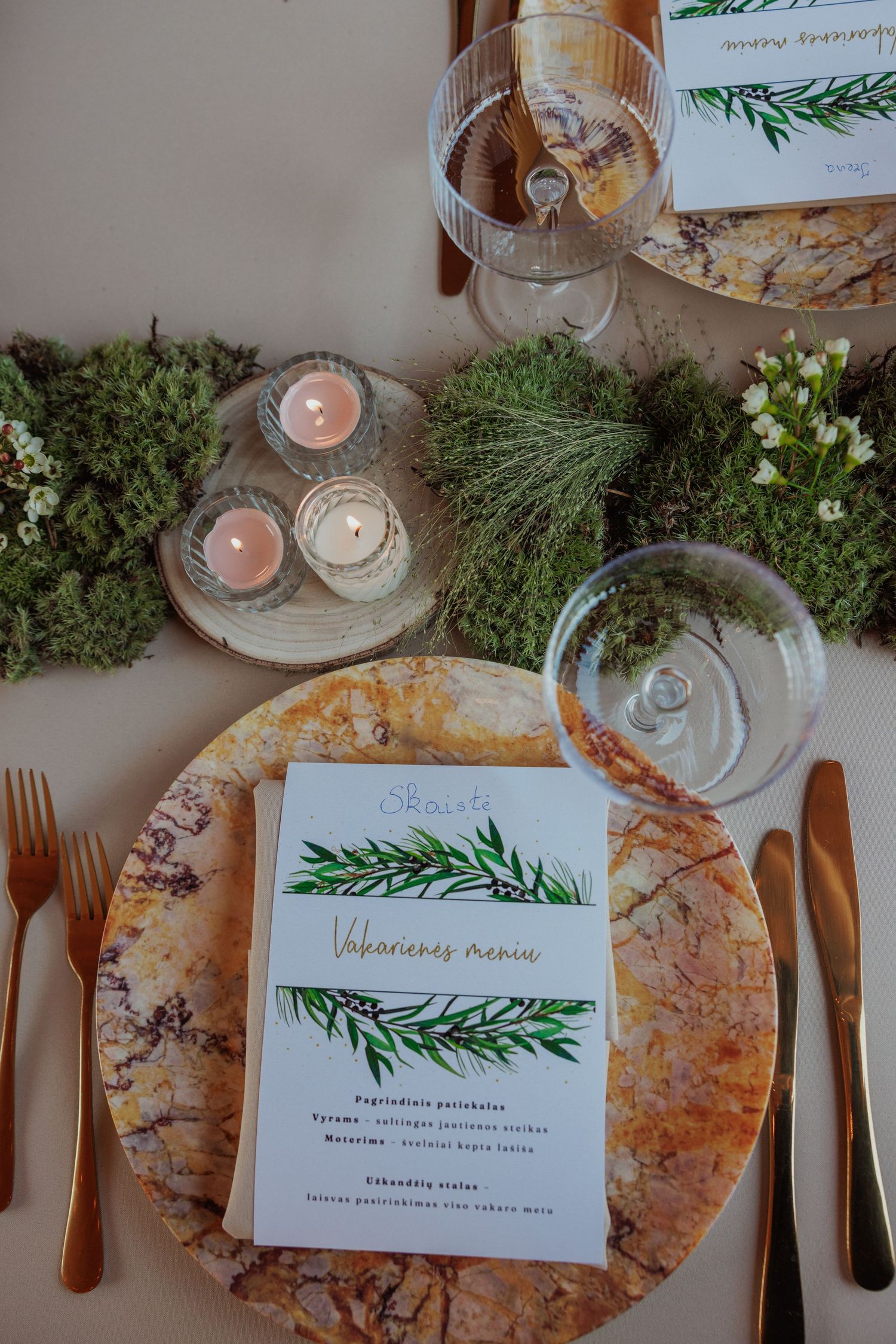 Elegant table setting with rosemary menu card, marble plates, and candles surrounded by greenery.