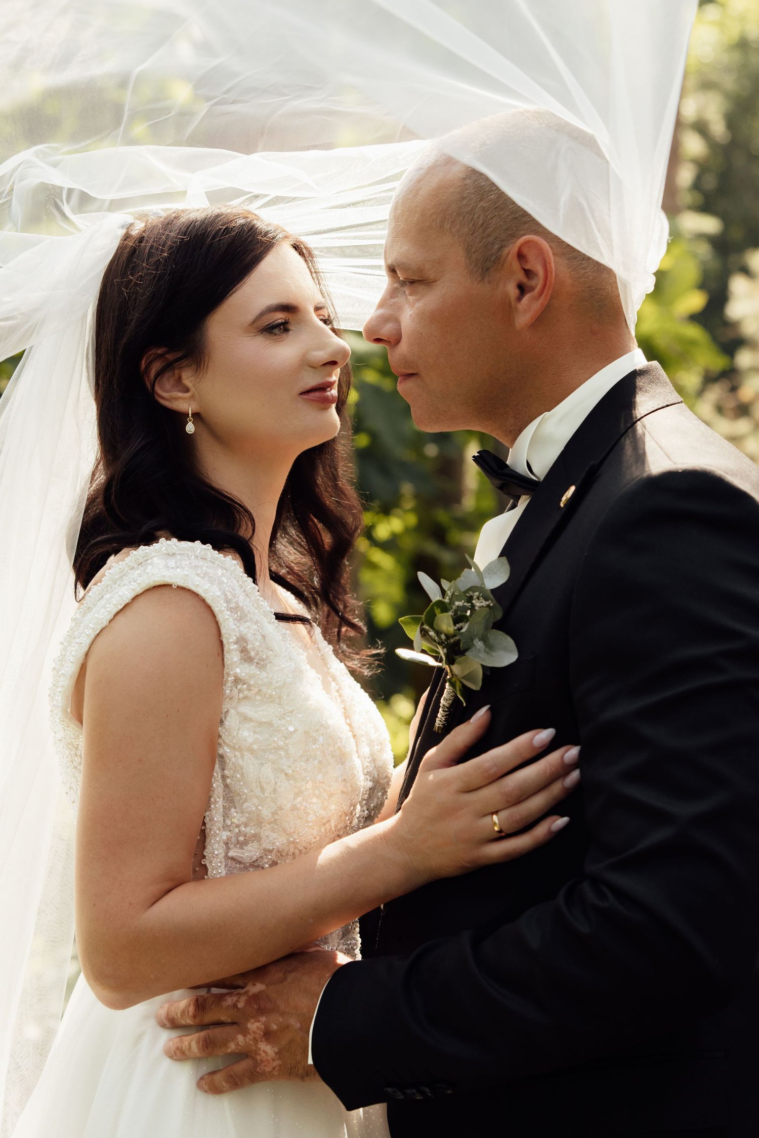 Bride and groom share a romantic moment under a flowing veil outdoors, dressed in wedding attire.