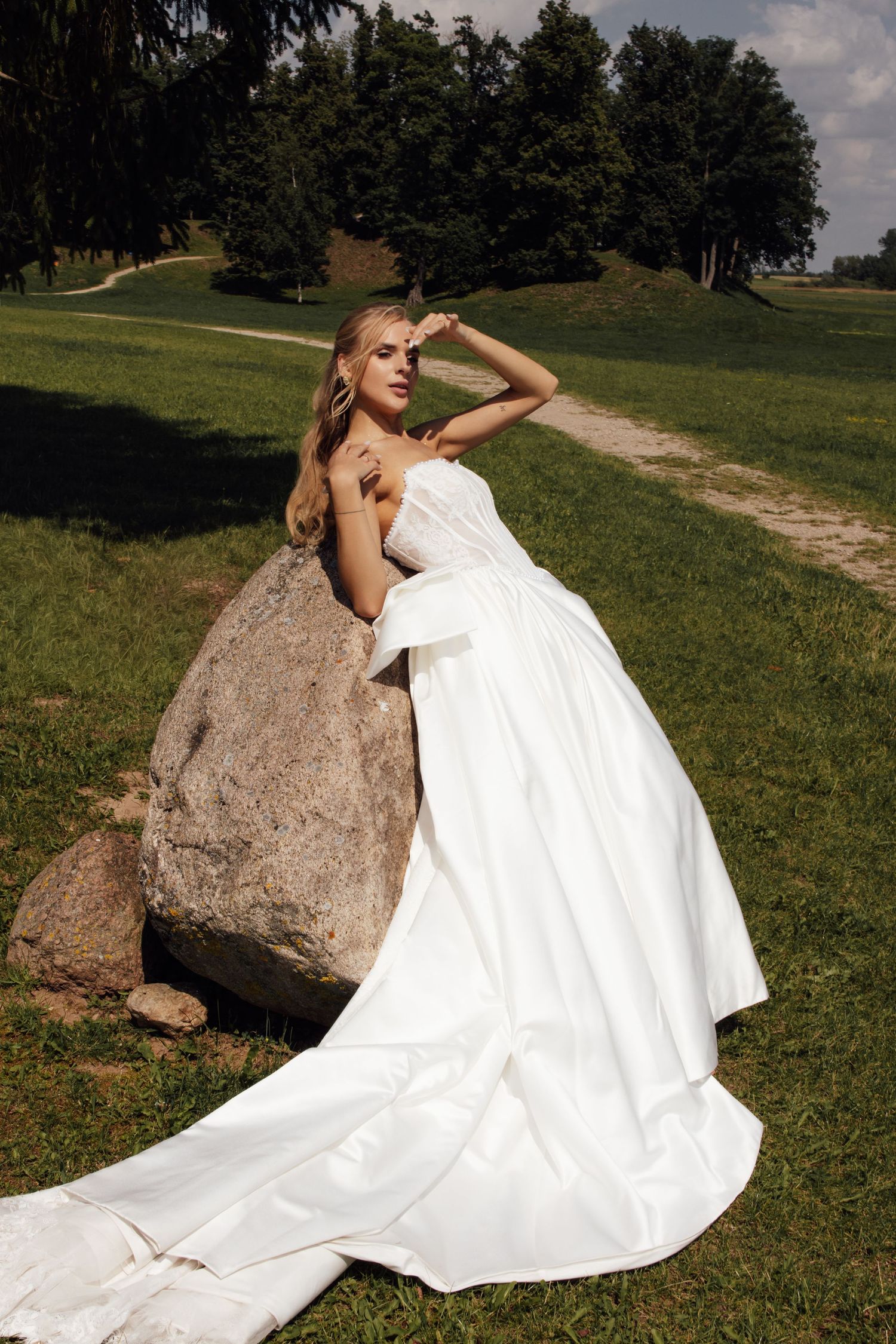 Elegant bride in a flowing white strapless ball gown posing against a large rock in a scenic outdoor setting.