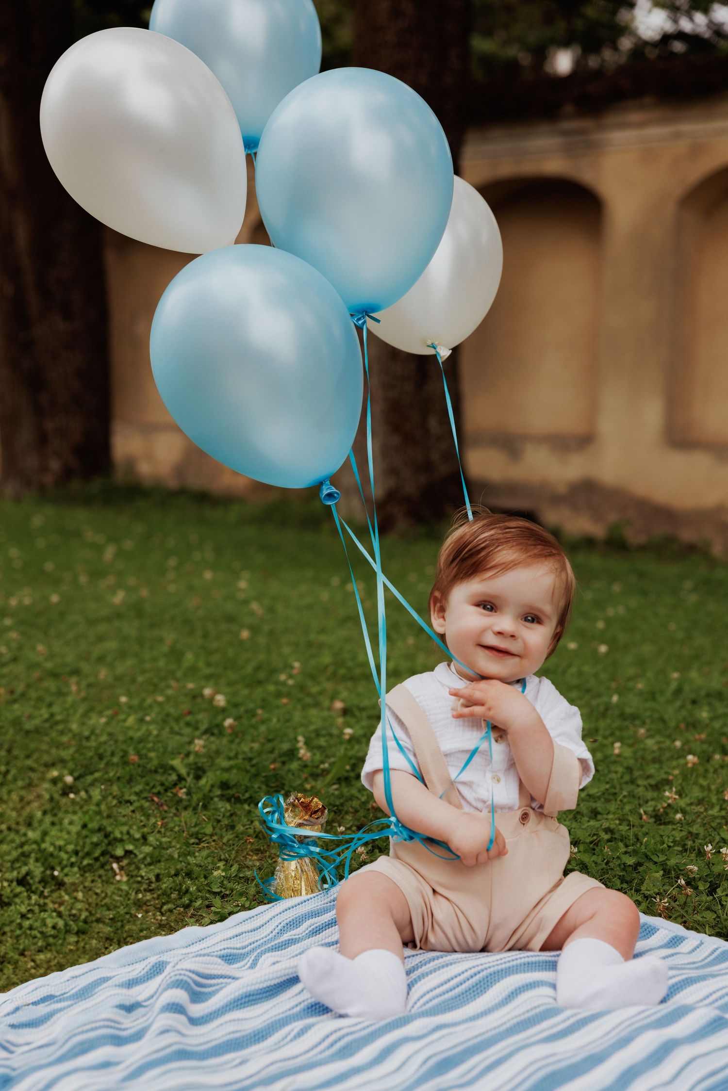 Baby sits on striped blanket in grass with blue and white balloons during outdoor photo session.