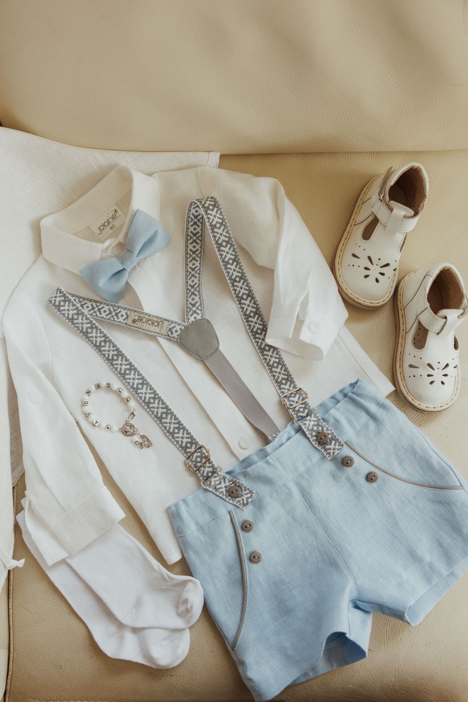 Baby formal wear set featuring white shirt, light blue vest, and white shoes laid out on beige surface.