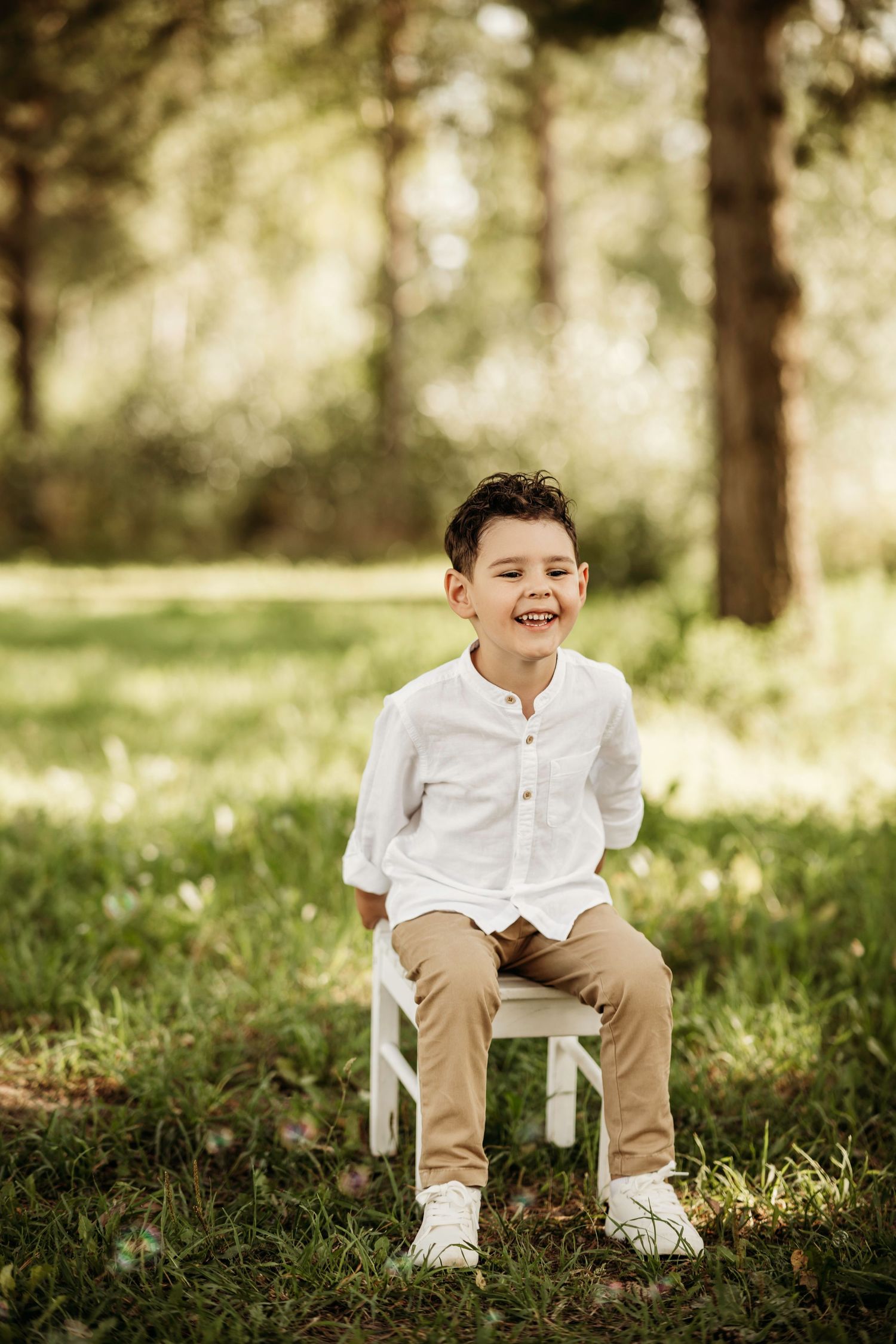 Child in white button-down shirt and khaki pants sitting on white chair outdoors in sunlit forest.