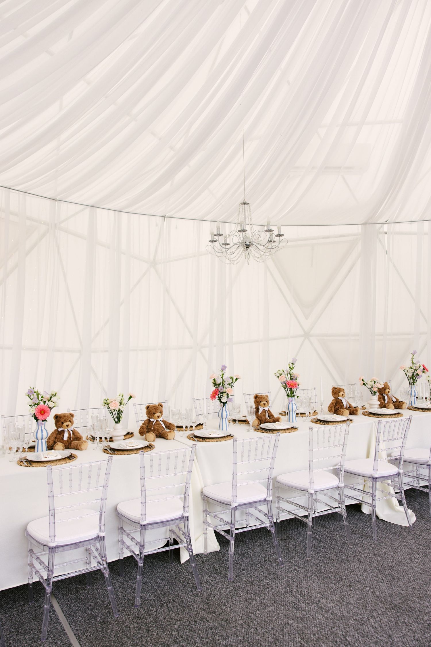 White tent reception setup with ghost chairs and teddy bear decorations on long tables.