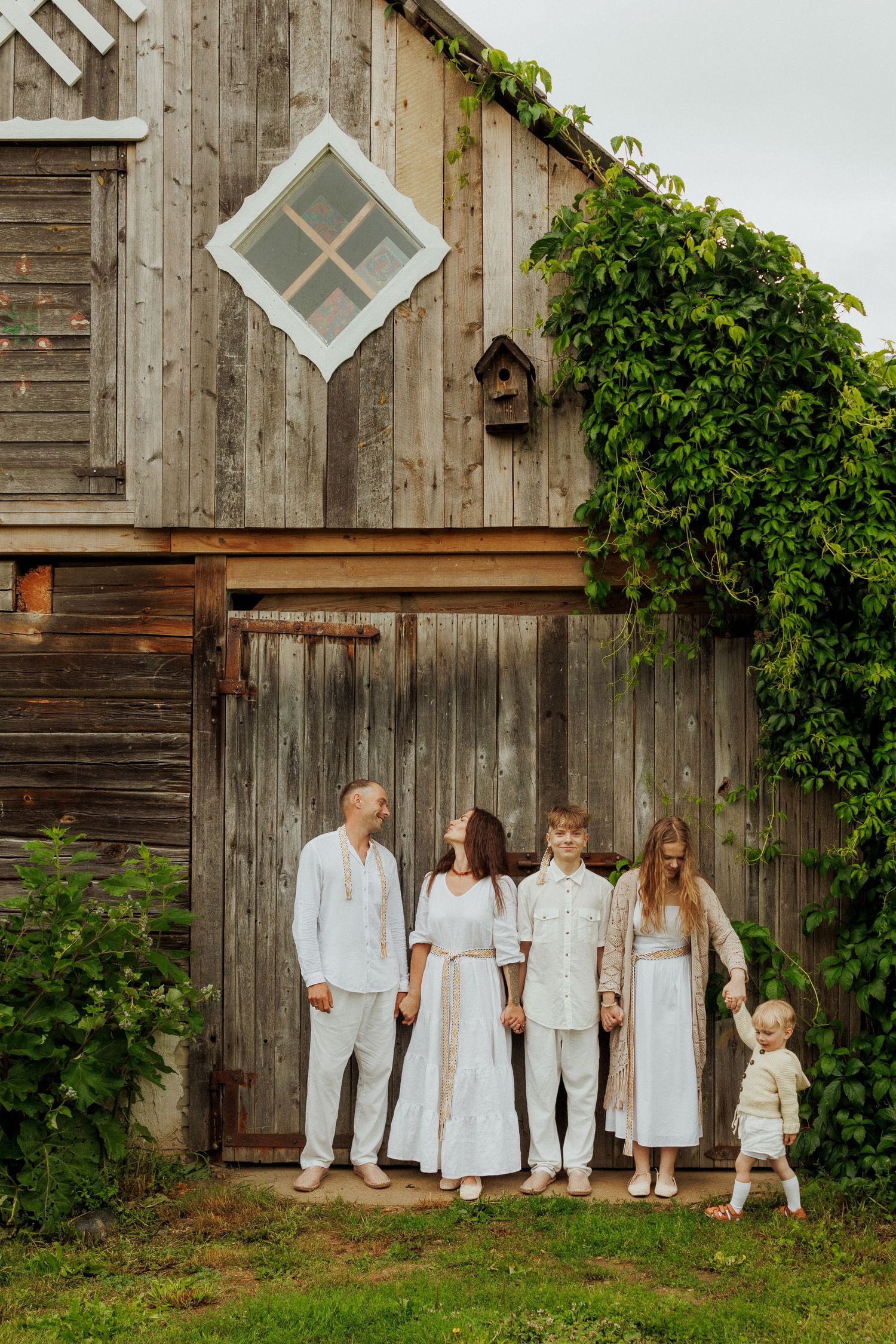 Family of five in white linen outfits standing before a rustic wooden barn covered in green ivy.