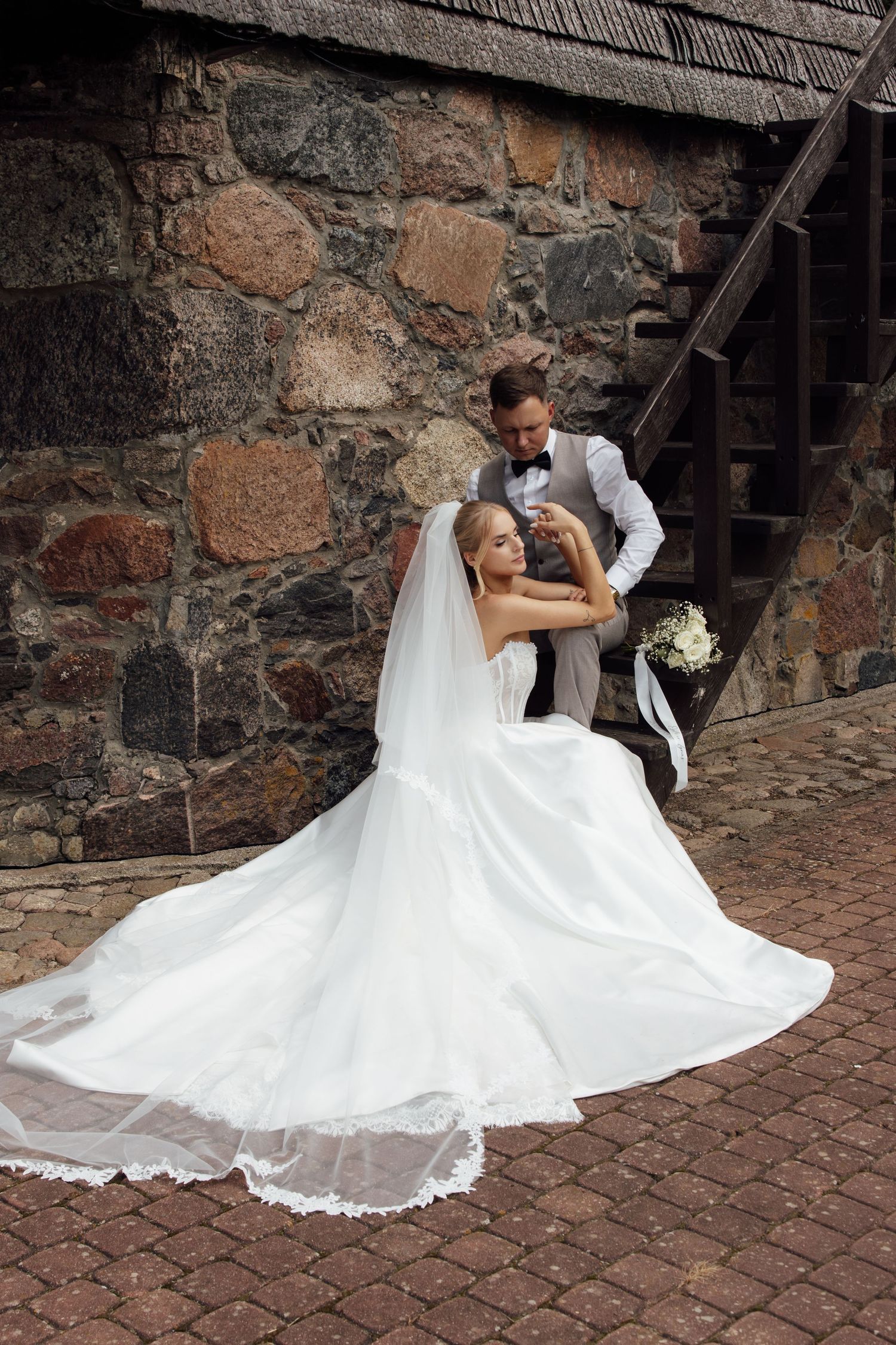Bride in white gown and veil poses with groom in gray suit near rustic stone wall on cobblestone path.