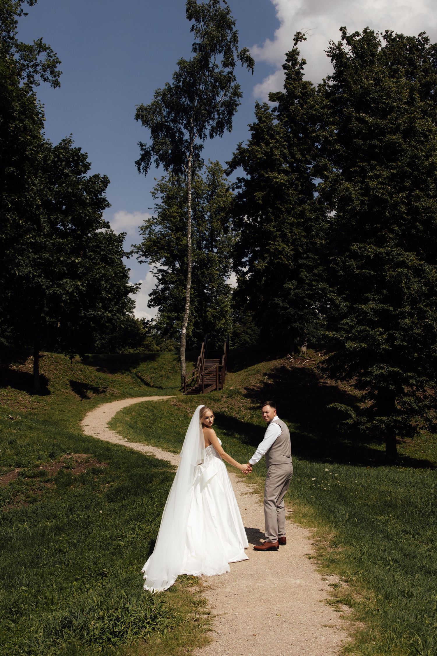 A bride and groom hold hands on a winding path surrounded by lush green trees under a blue sky.