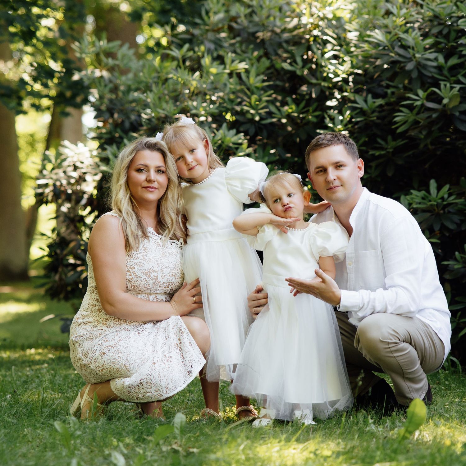 A happy family of four poses outdoors on green grass, parents in white outfits with two young daughters in white dresses.