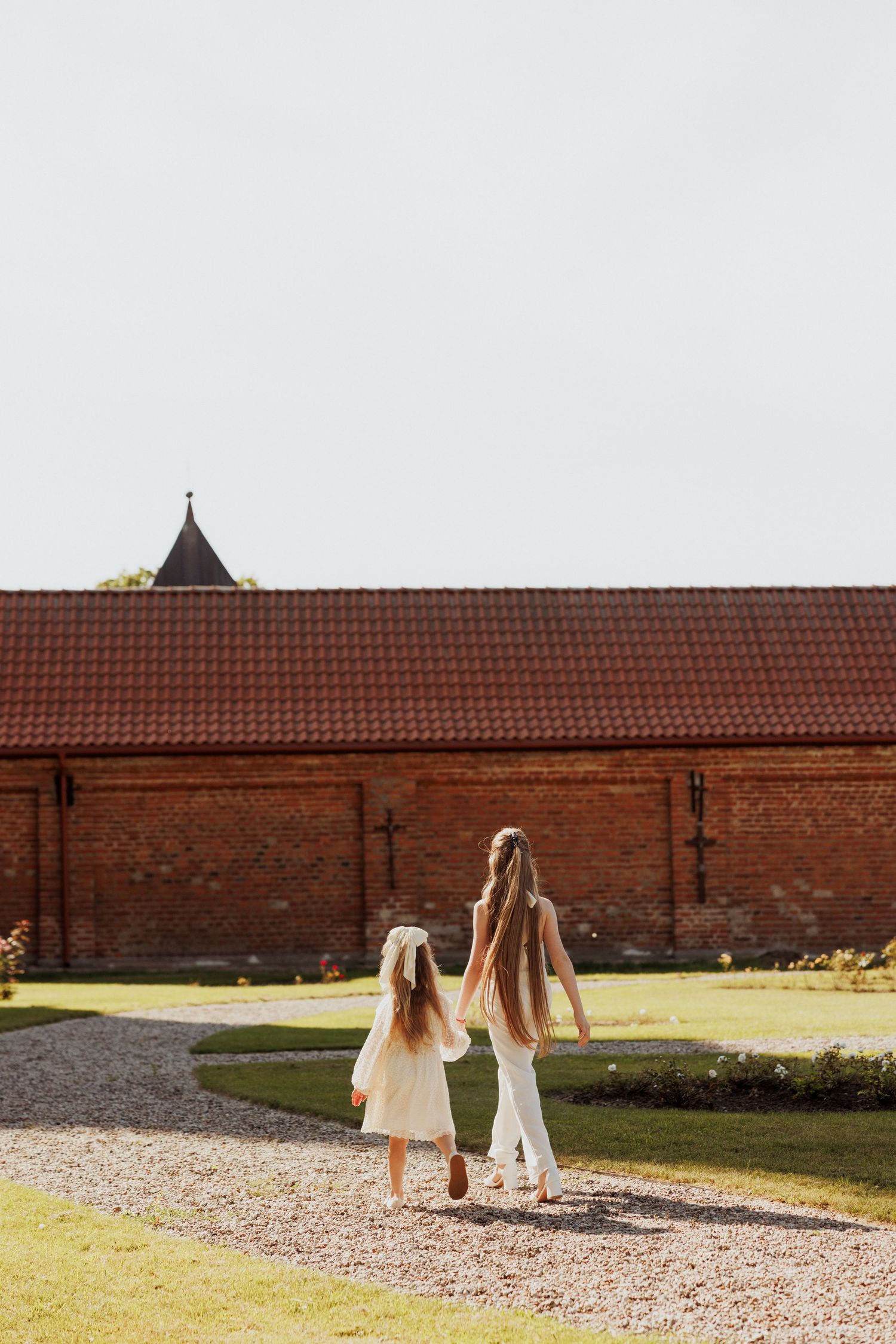 Two people walking on a gravel path alongside a historic brick wall on a sunny day.