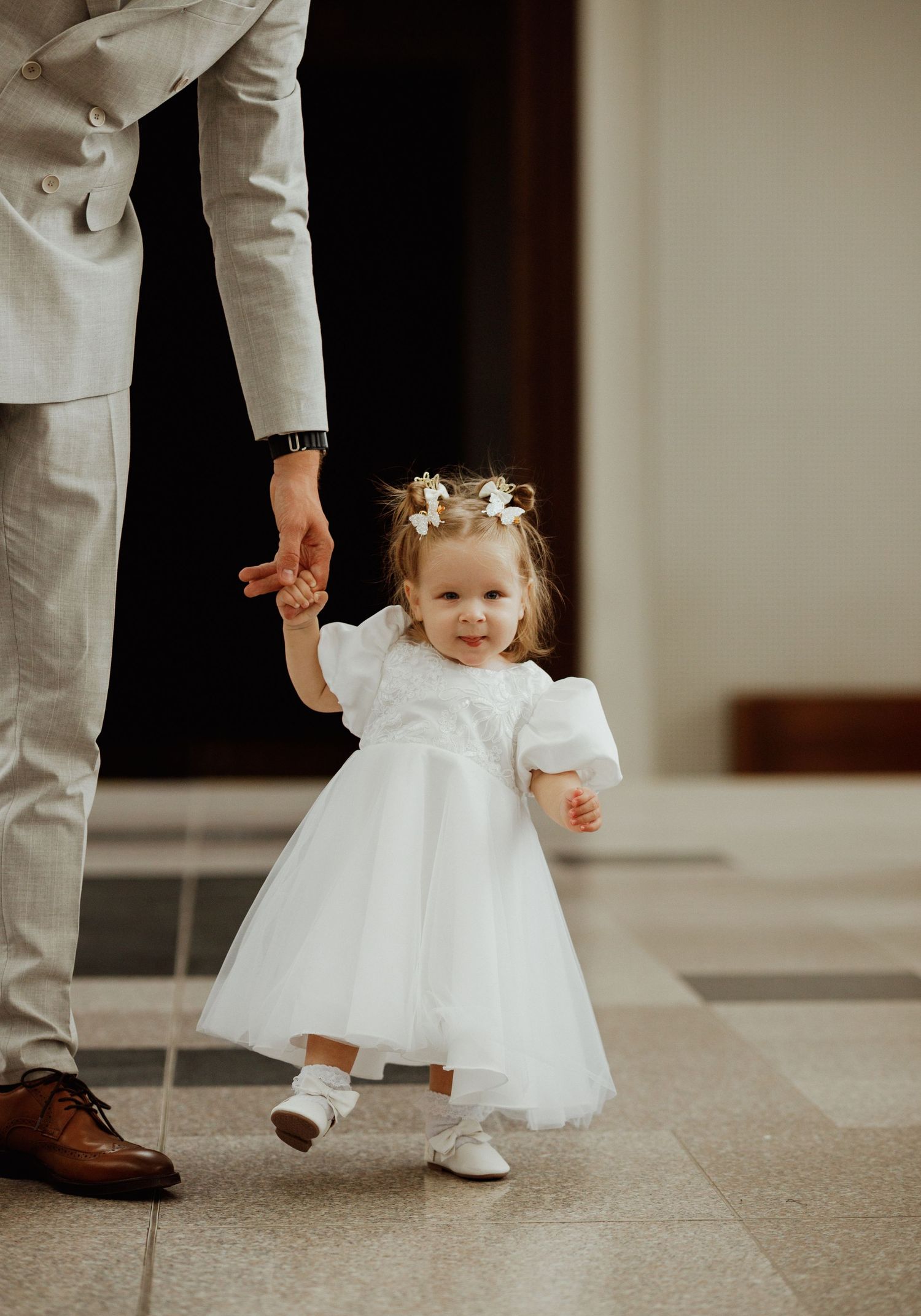 A small child in a white dress stands and dances with an adult wearing white pants and brown shoes.