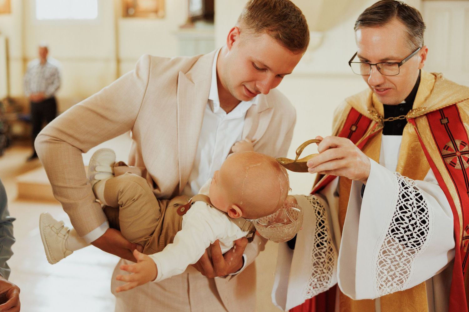 A baby receives a blessing during a religious baptism ceremony in a church while wearing white christening attire.