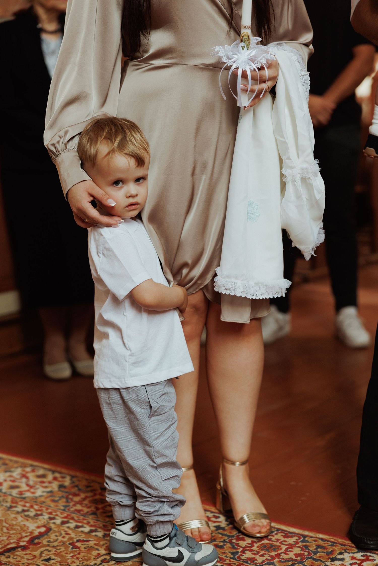 A small child in white shirt and gray pants holds onto someone's dress during a formal event.