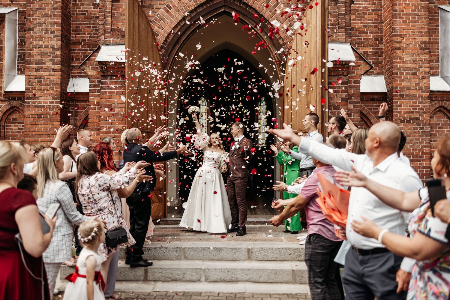 Wedding guests throw confetti over the newlyweds outside a historic brick church building.