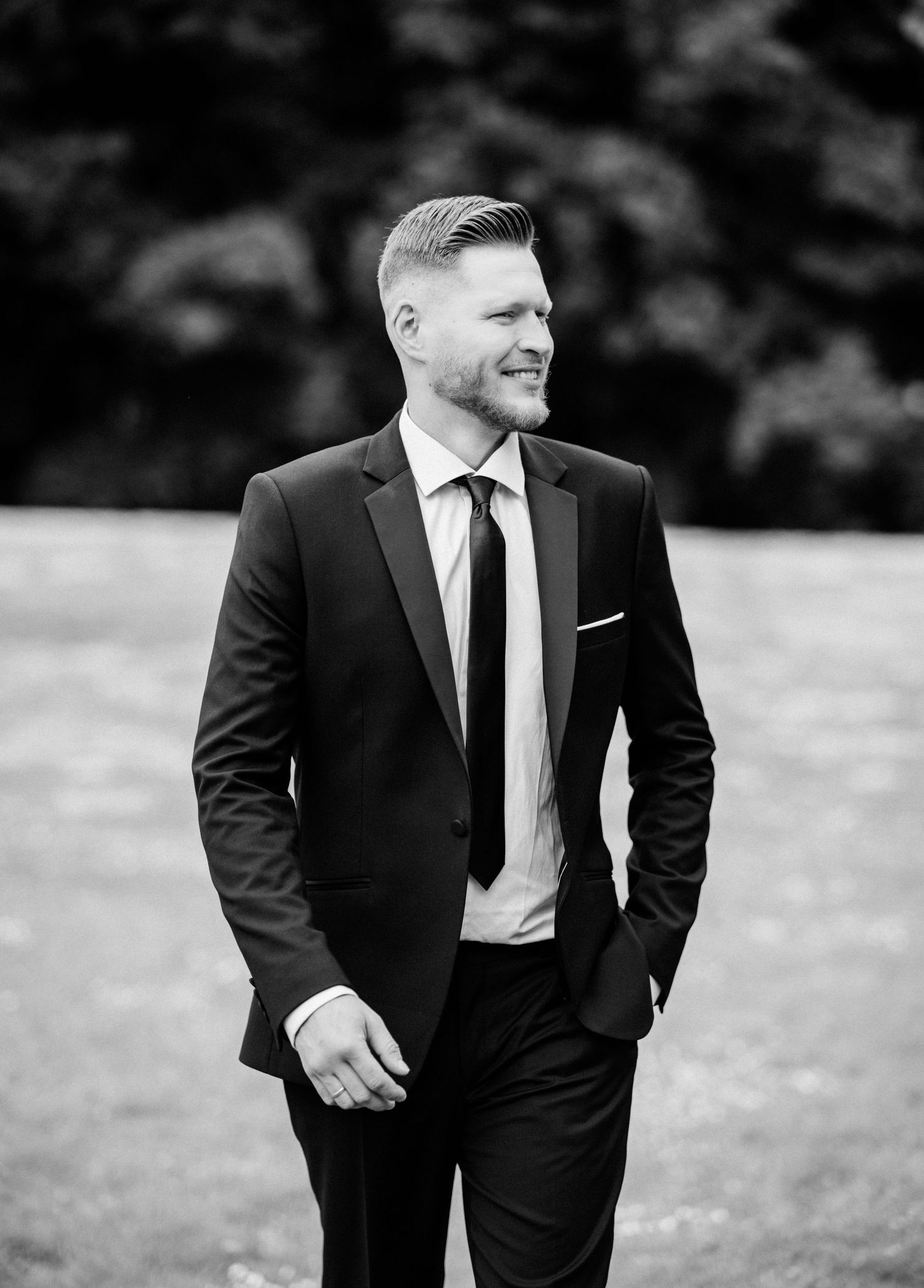 A black and white portrait of someone in a well-fitted suit and tie smiling outdoors with a blurred background.