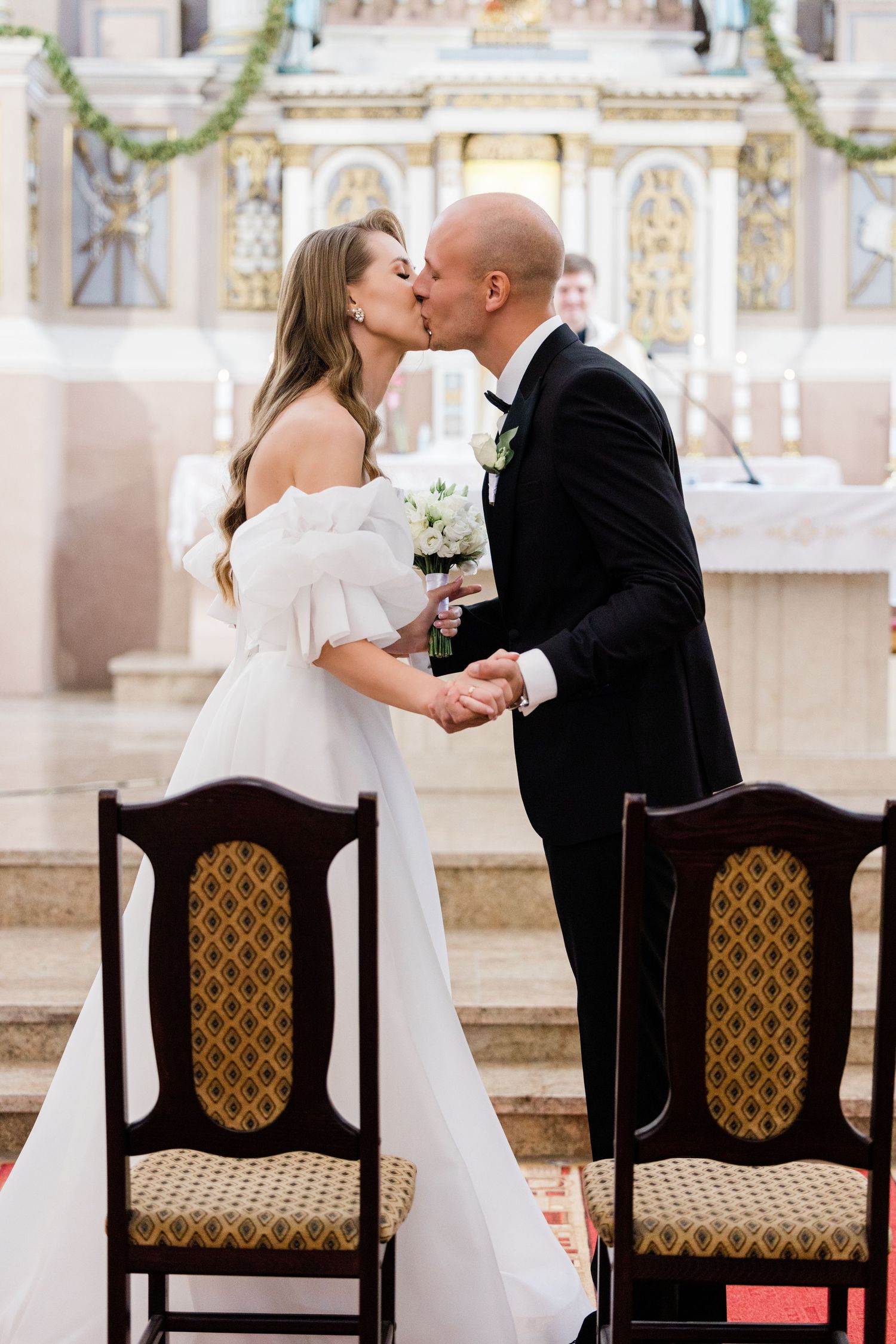 Romantic wedding ceremony moment showing couple seated in ornate chairs with decorative upholstery in church setting.