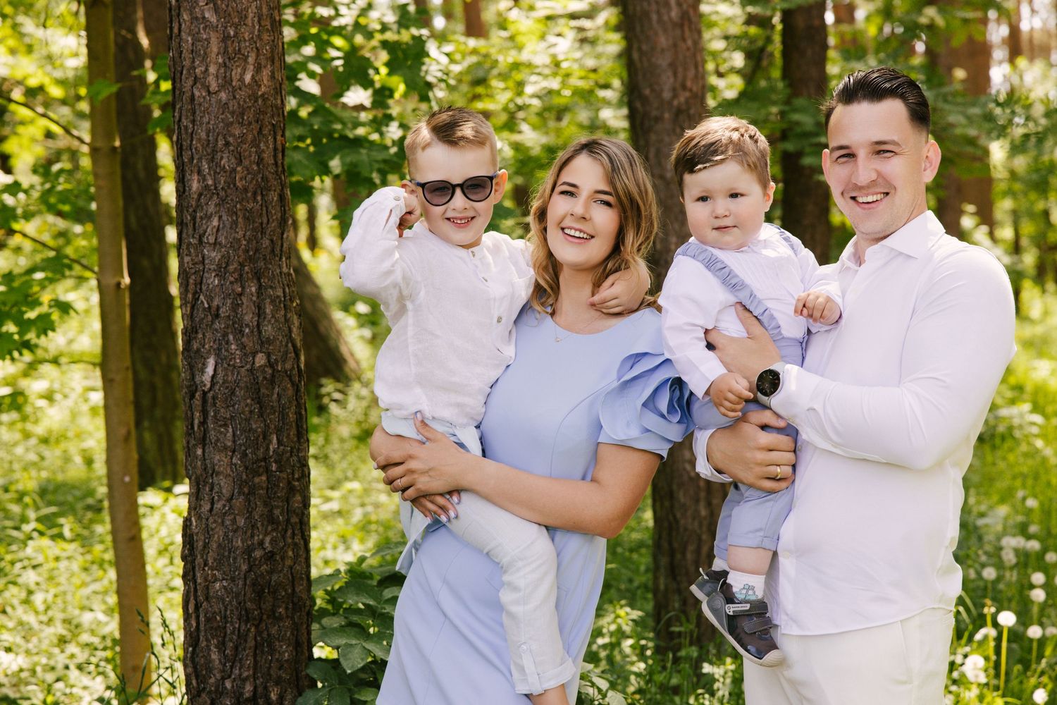Family wearing coordinated white and blue outfits poses together in a sunlit forest setting.