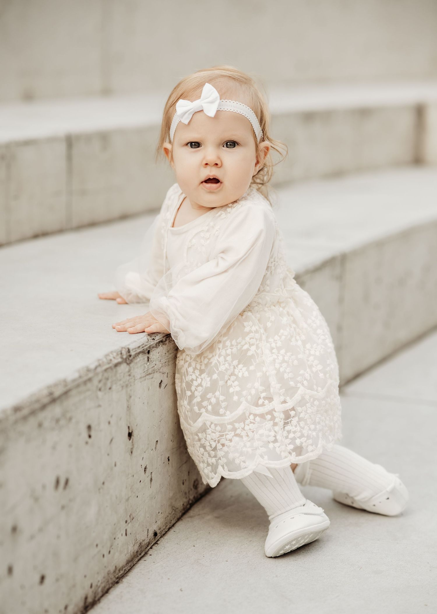 Small child in white lace dress and bow sitting on concrete steps in natural lighting.