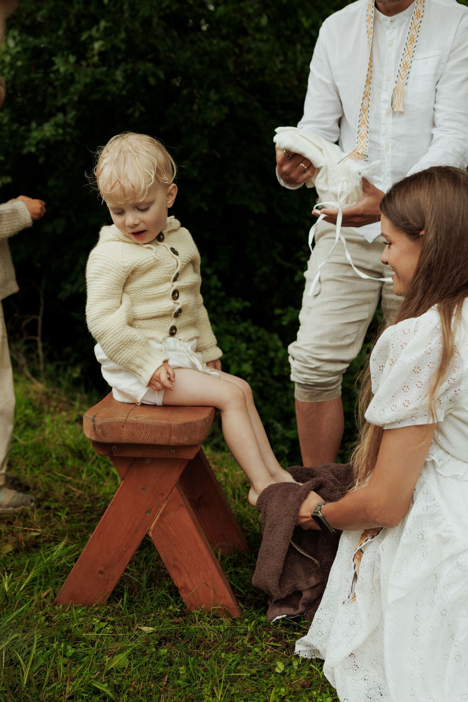 Young child sitting on red wooden stool while girl in white dress helps put on shoes outdoors.