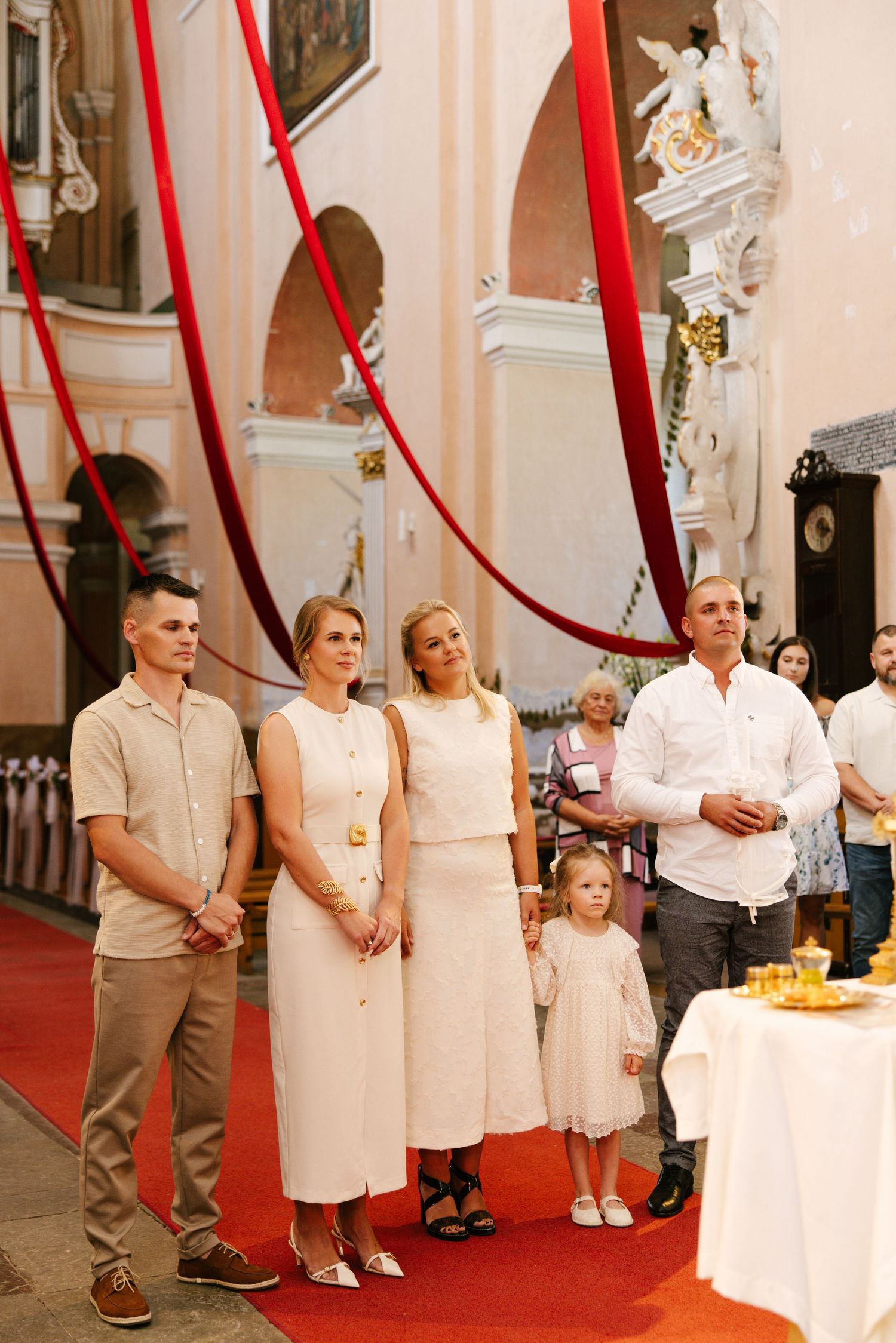 A group stands in formal white attire along a red carpet in an ornate church interior.