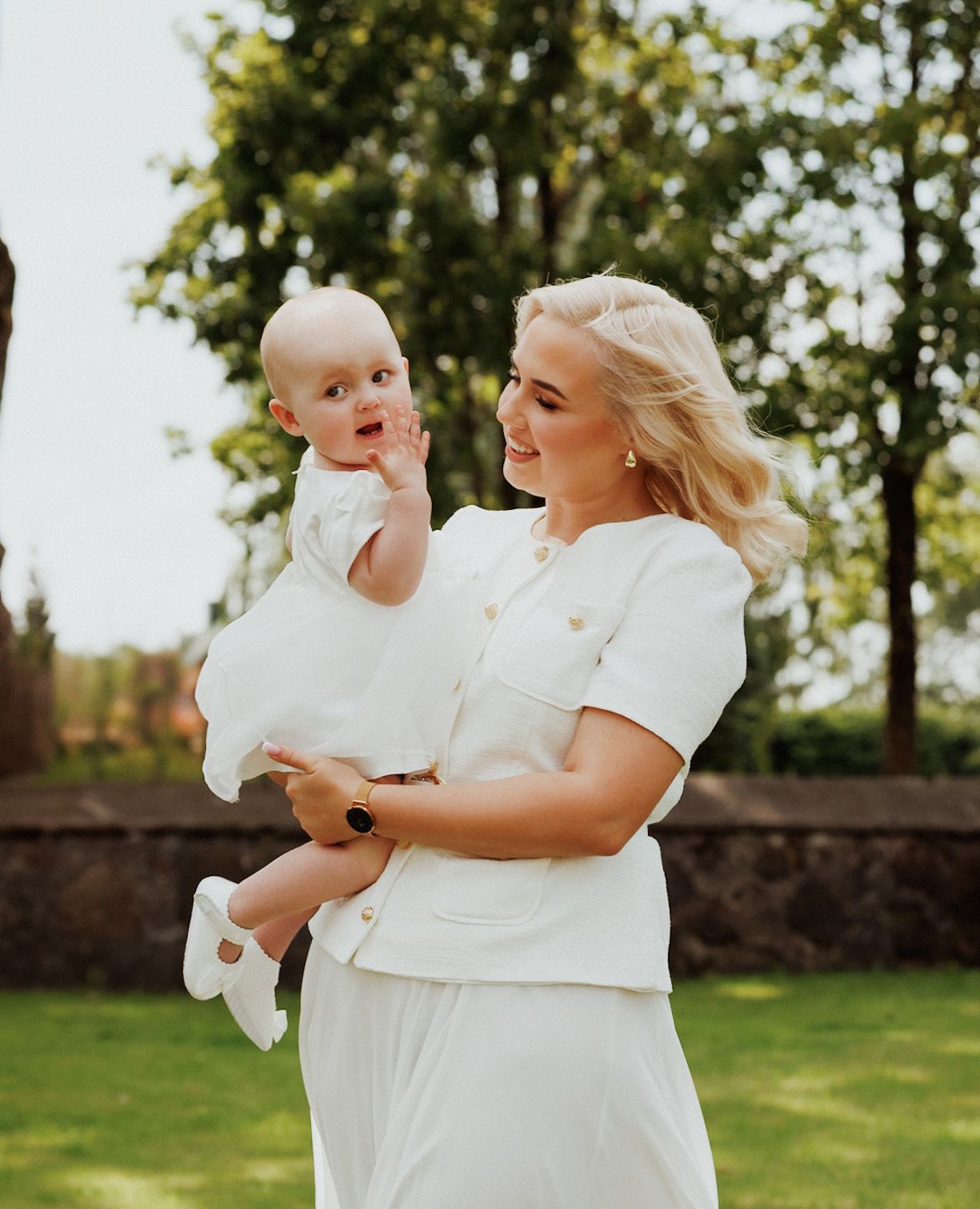 Mother and baby in matching white dresses share playful moments on a sunny lawn with trees in background.