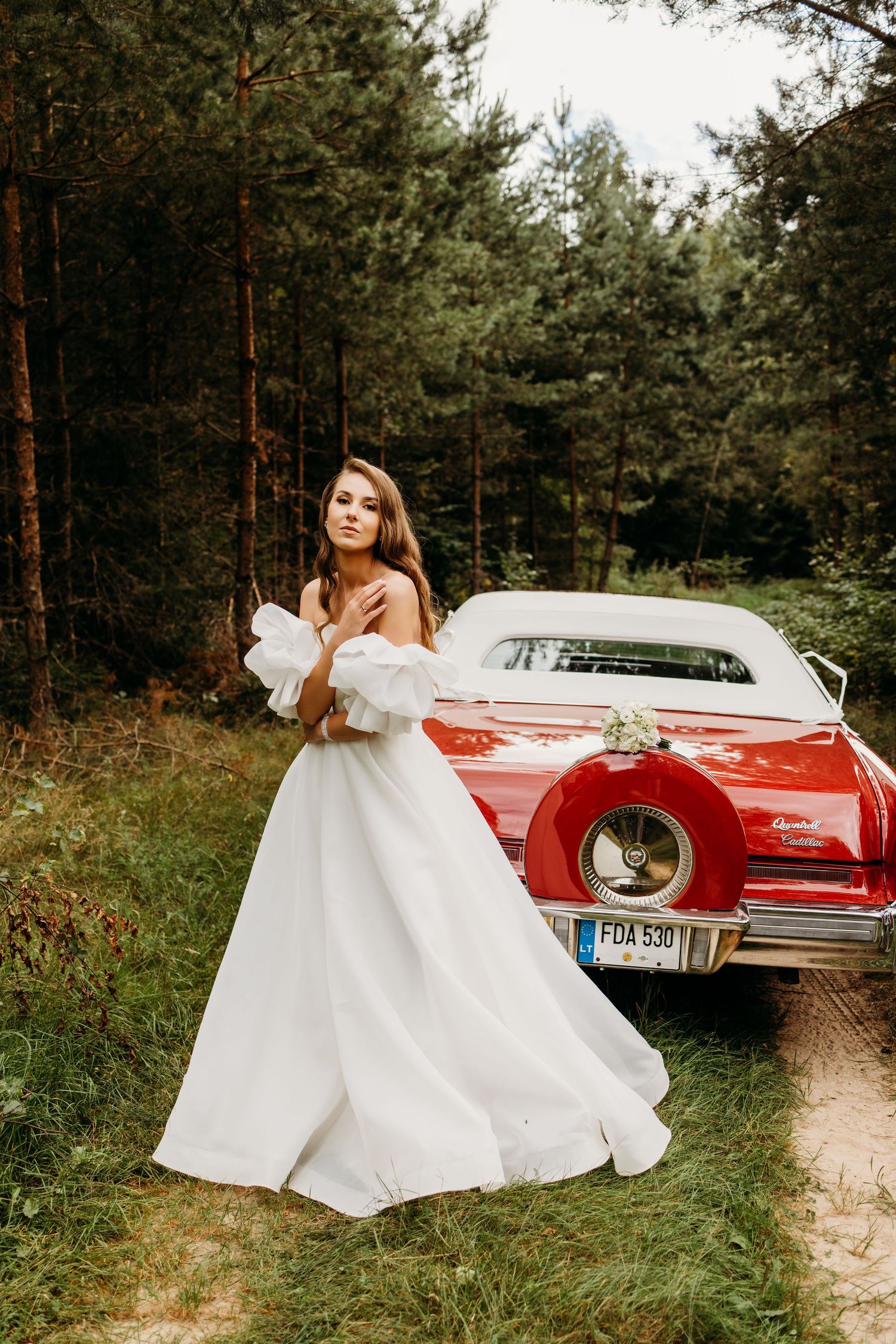 A bride in an elegant white off-shoulder gown poses in front of a classic red car in a pine forest setting.