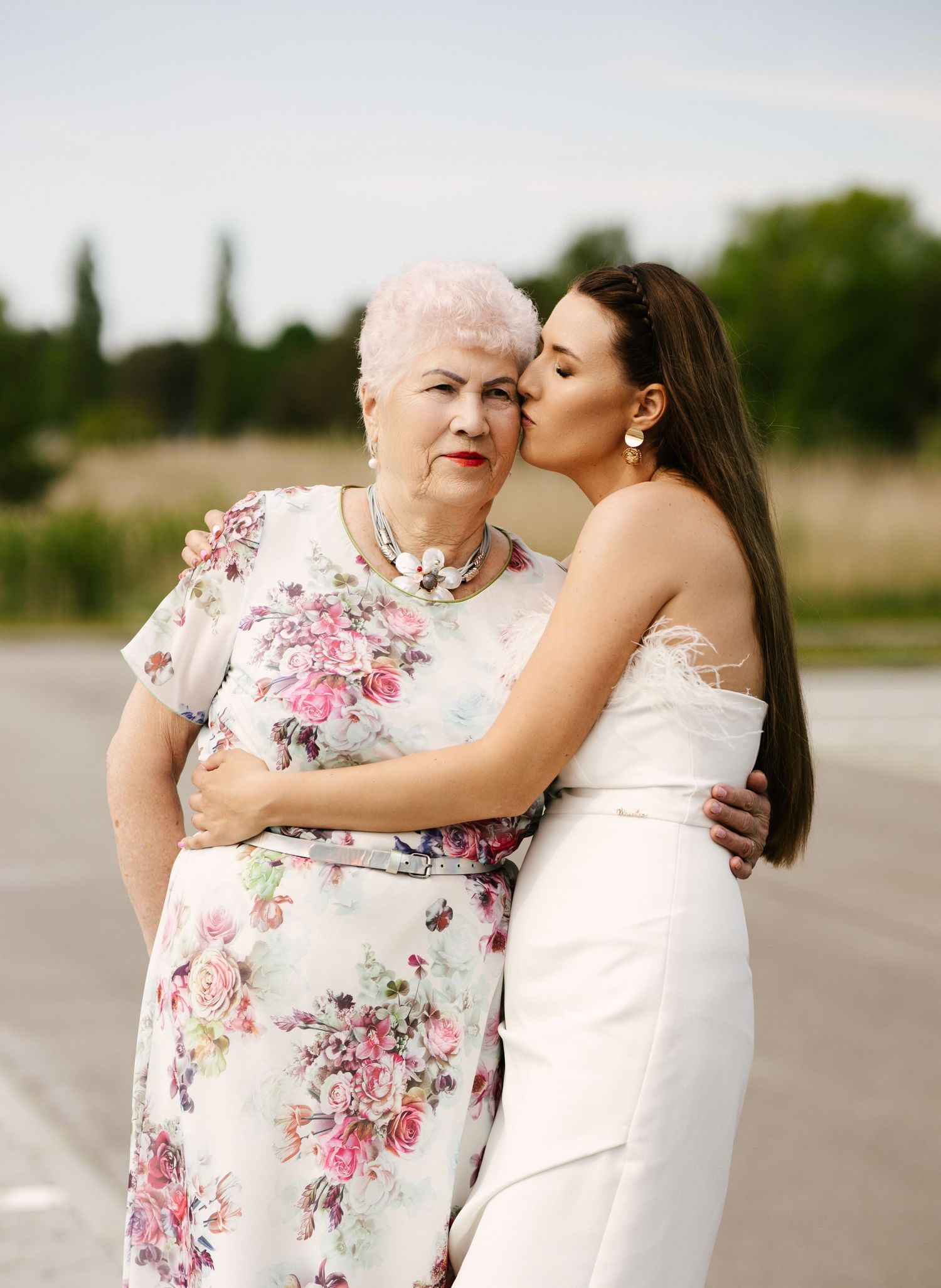 A woman in a white dress embraces and kisses someone wearing a pink floral dress in a heartwarming outdoor moment.