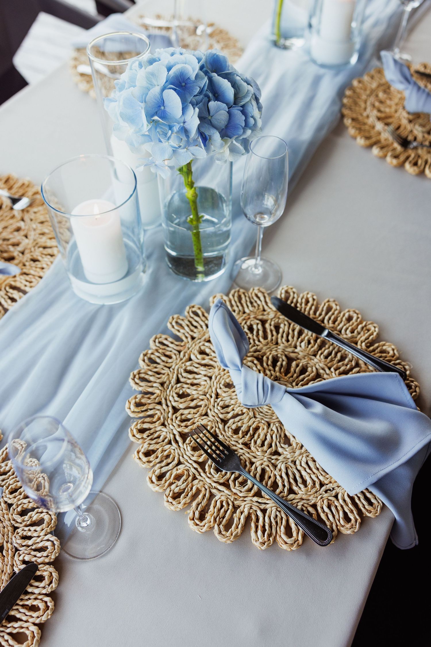 Elegant table setting with light blue hydrangeas, white candles, and gold woven placemats creating a luxurious atmosphere.
